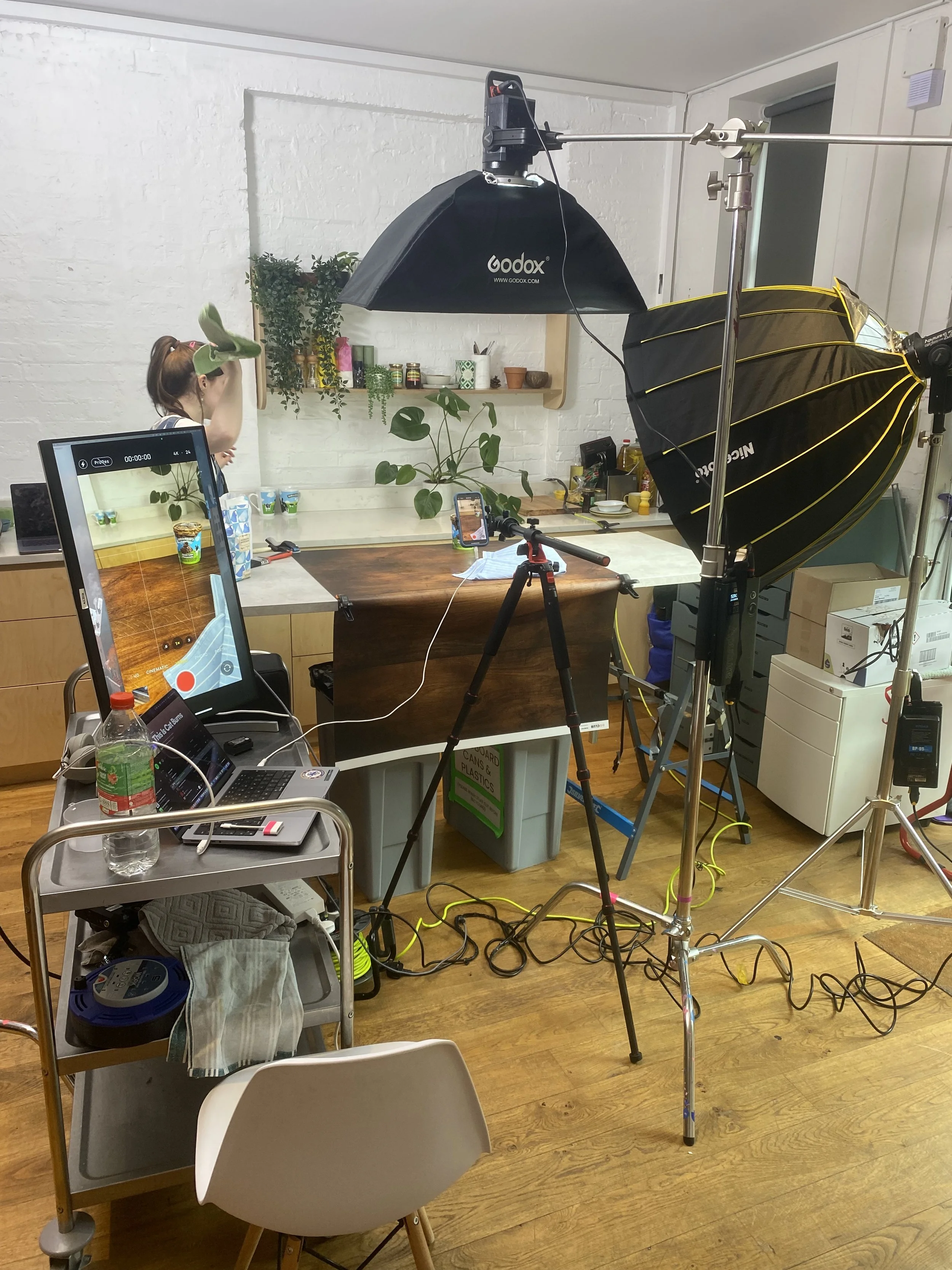 A photography studio setup in a room with a white brick wall and wooden floor, including professional lighting equipment, a camera on a tripod, a computer monitor, and a person adjusting her hair in the background.