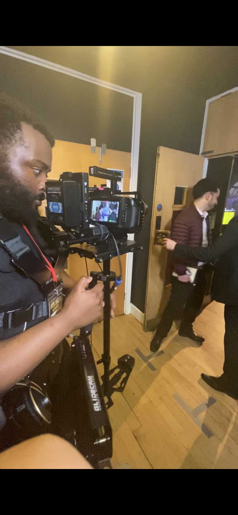 A film crew member manages camera equipment while filming indoors, with people in the background near a door and a television.