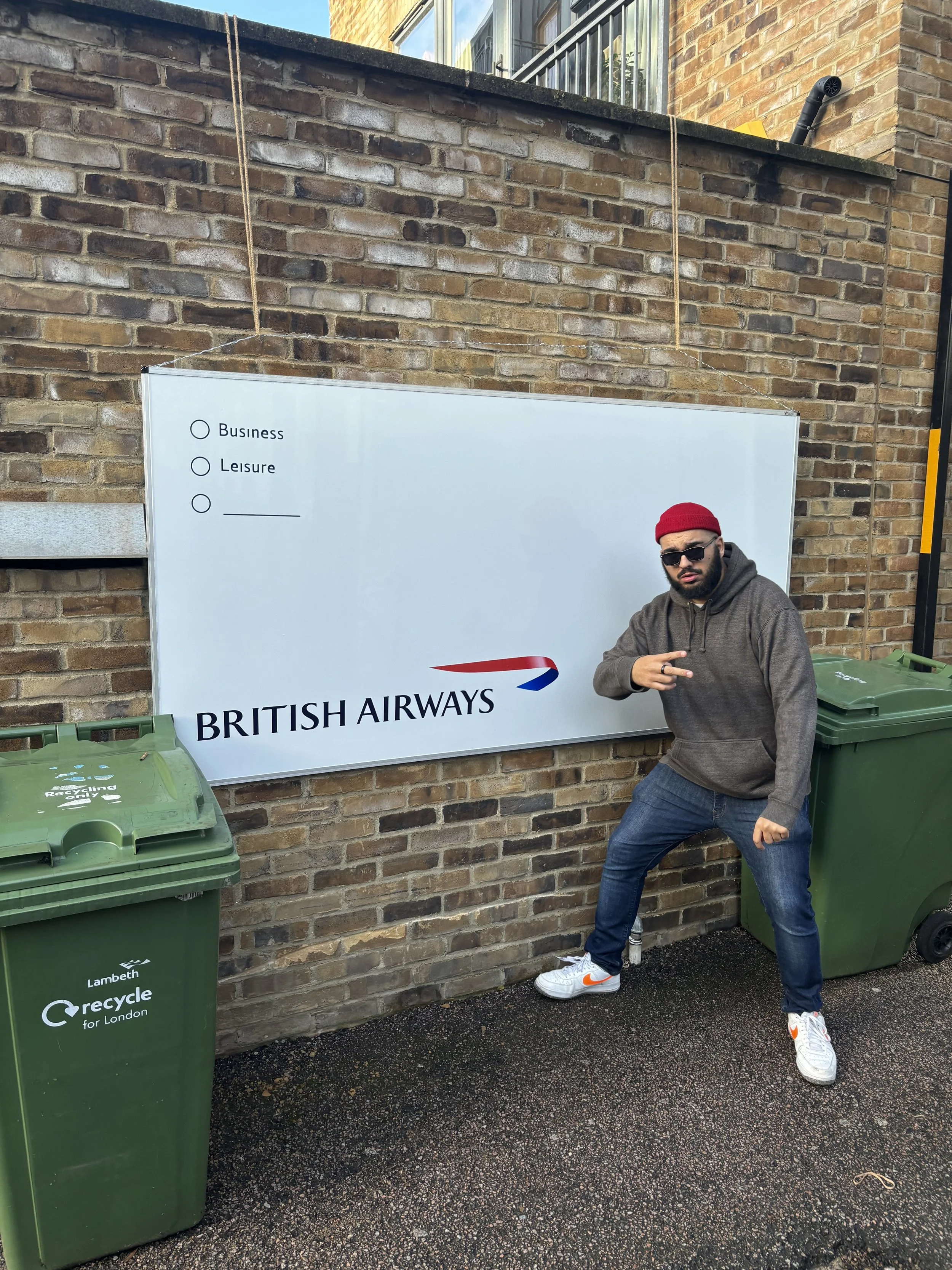 A man wearing sunglasses, a red beanie, a brown hoodie, and jeans standing next to a British Airways sign and two green recycle bins outside a brick building.