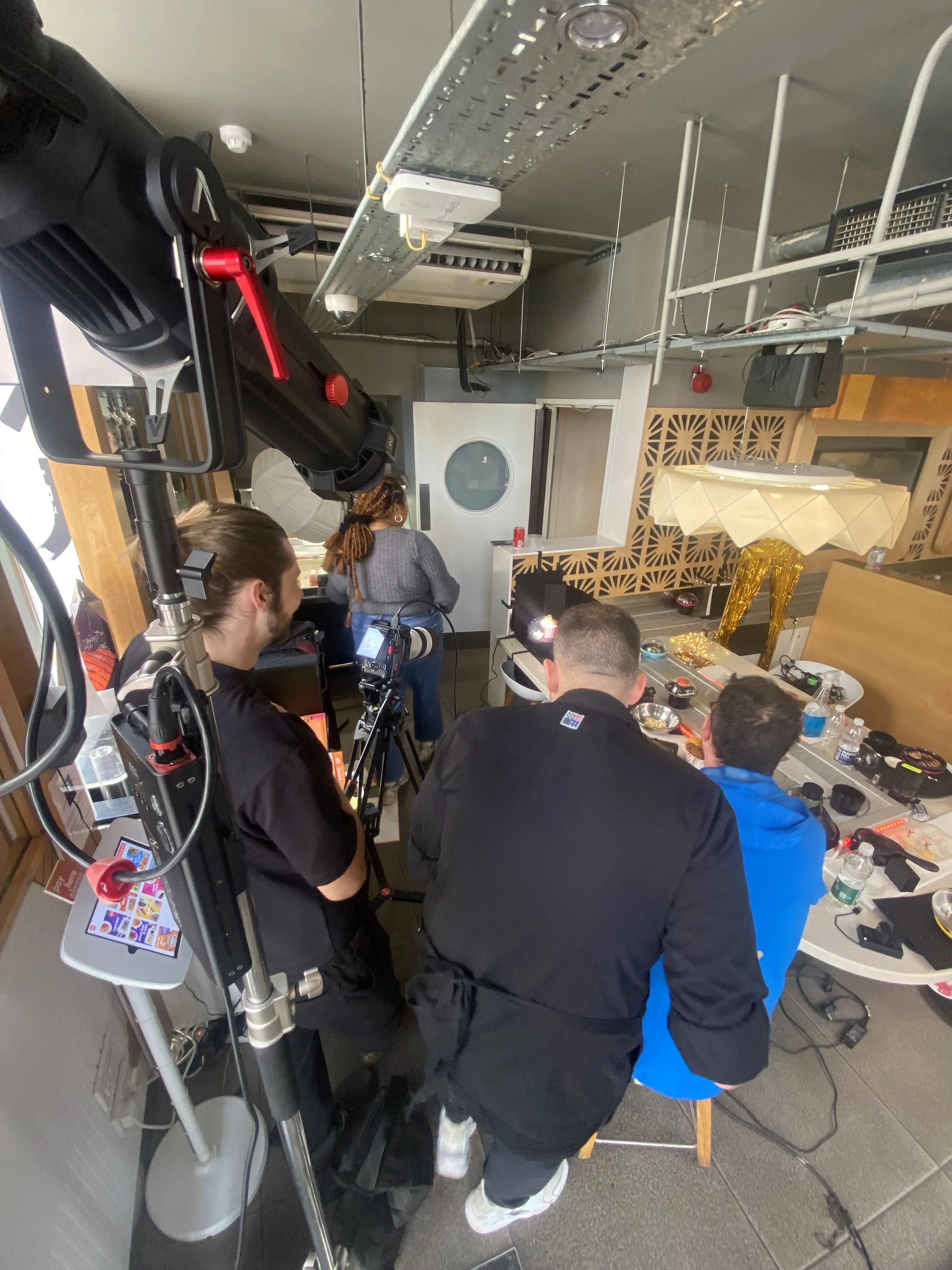 Photographers and videographers filming a live cooking show or demonstration in a restaurant or studio setting, with a table of ingredients and equipment visible.
