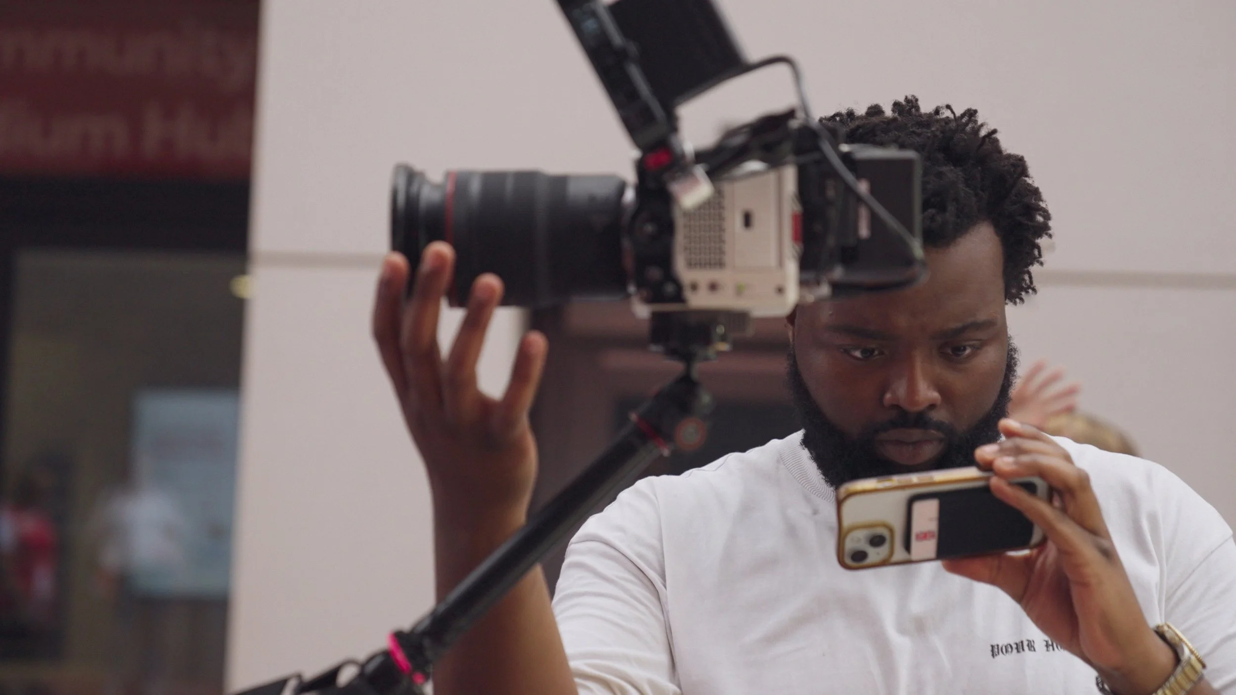 A man operating a professional video camera while looking at his smartphone, in an indoor setting.