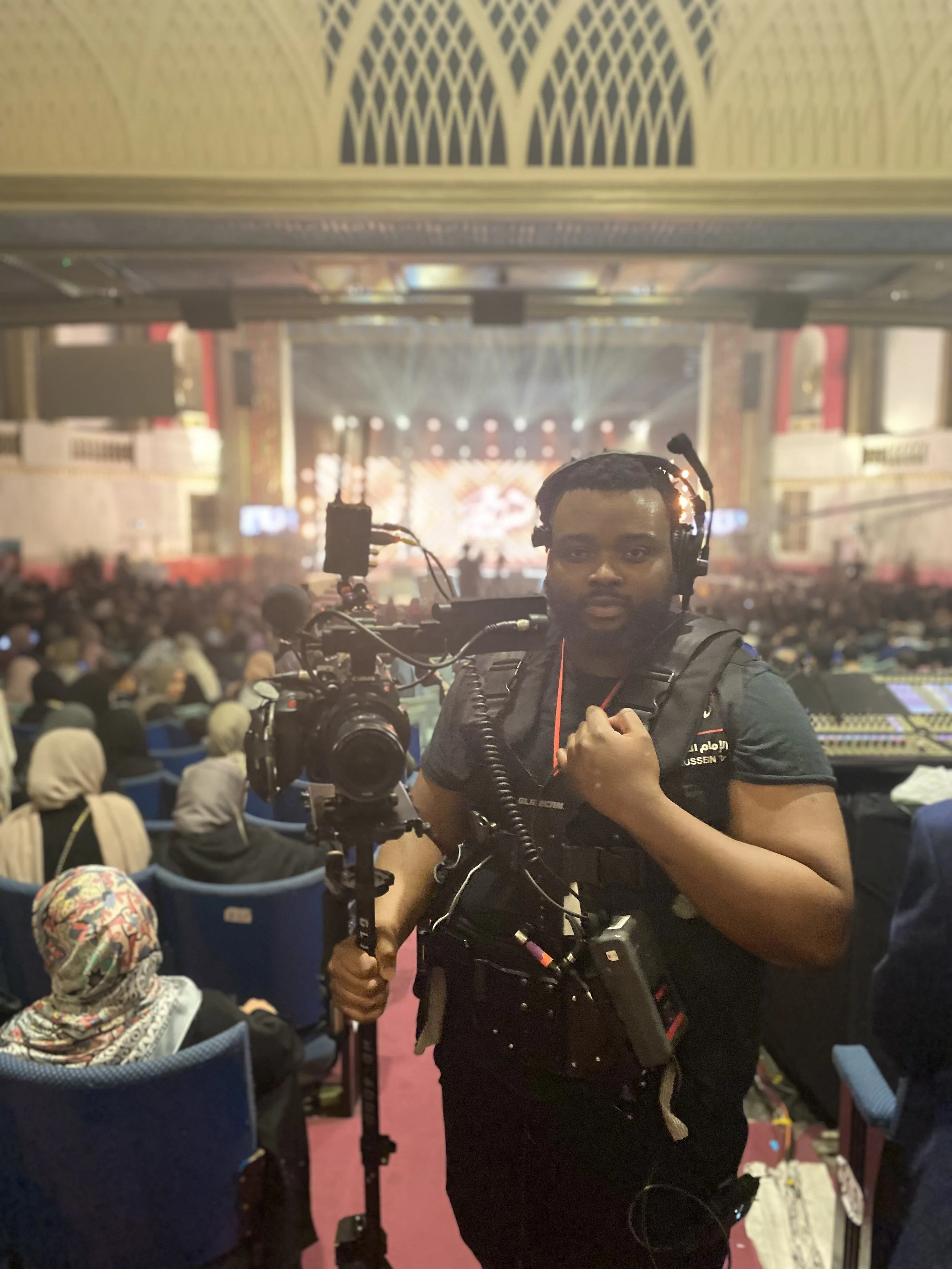 A man operating a professional camera at a large indoor event with a stage and bright lights in the background, surrounded by a seated audience.