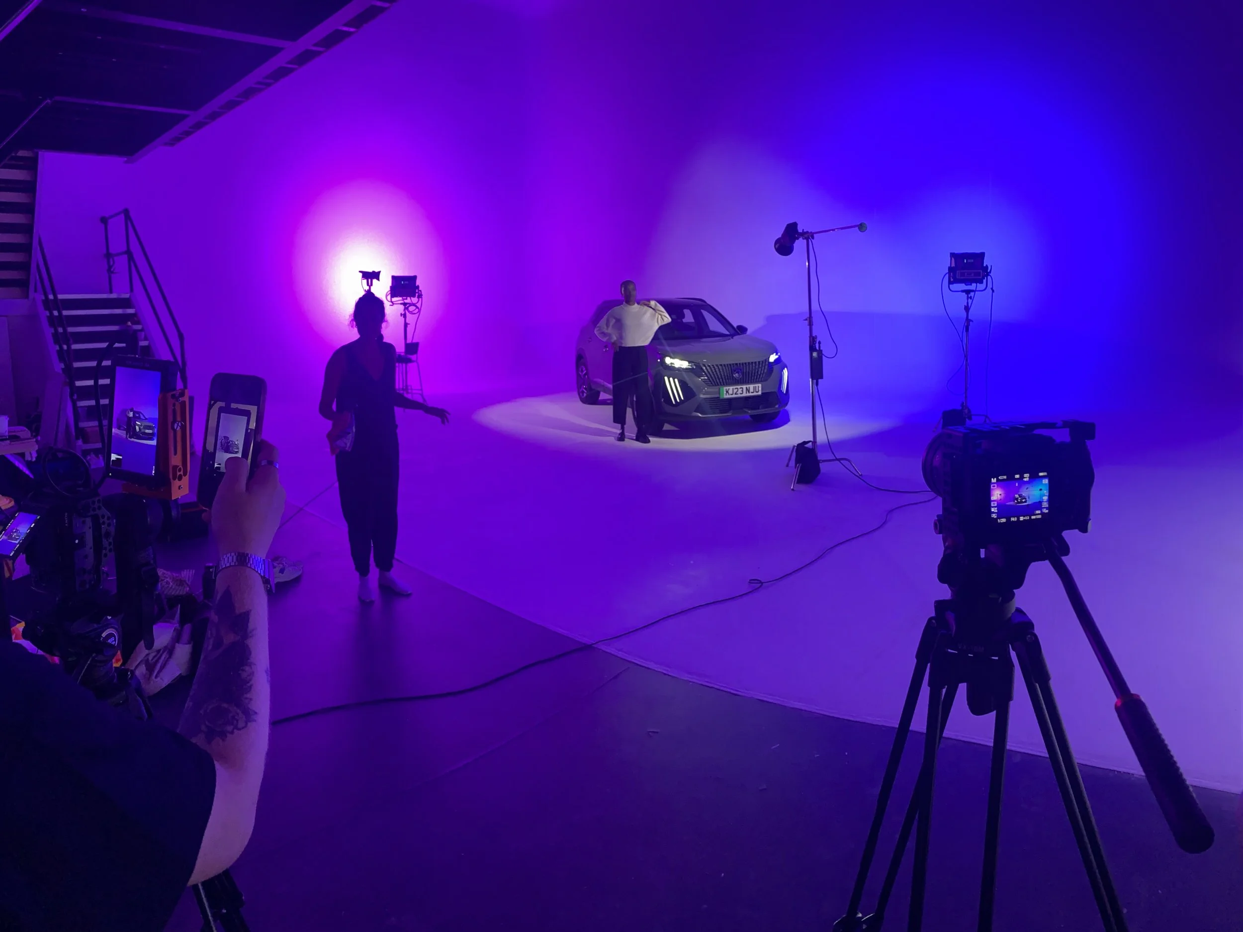 A photo of a photoshoot studio with a car under purple and blue lighting, with a woman posing and a man standing beside the car. Cameras and lights are set up around the scene.