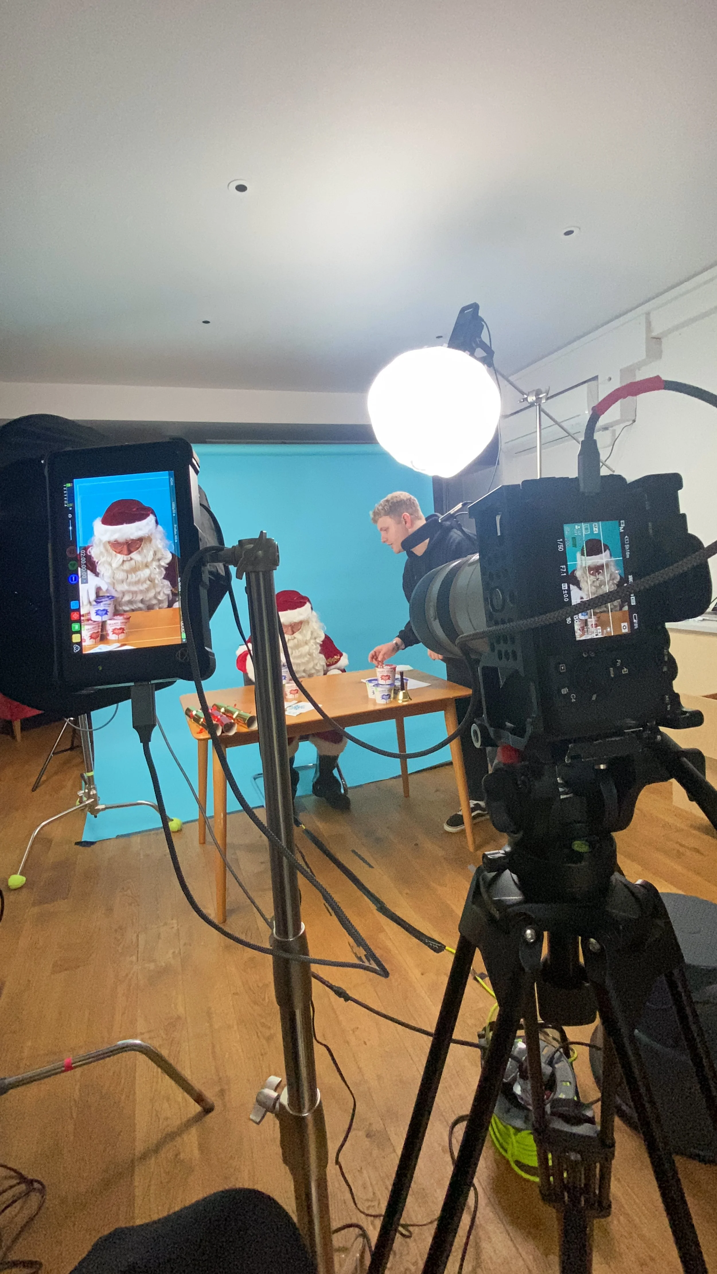 Behind the scenes of a video shoot featuring people dressed as Santa Claus and a person in black shirt, with camera equipment and studio lighting, in front of a blue backdrop.