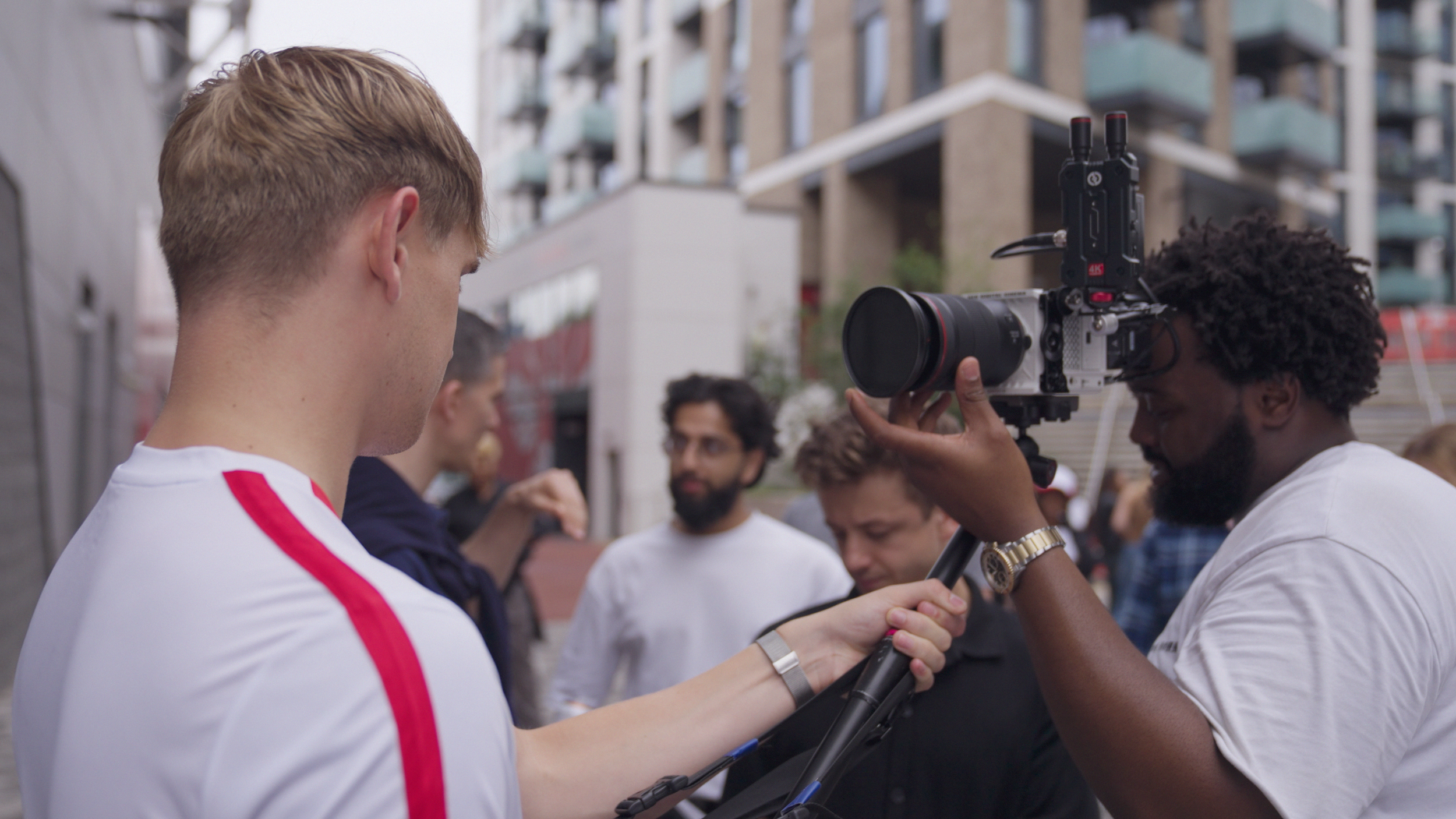 Two men are filming with a camera on a city street, surrounded by other people.