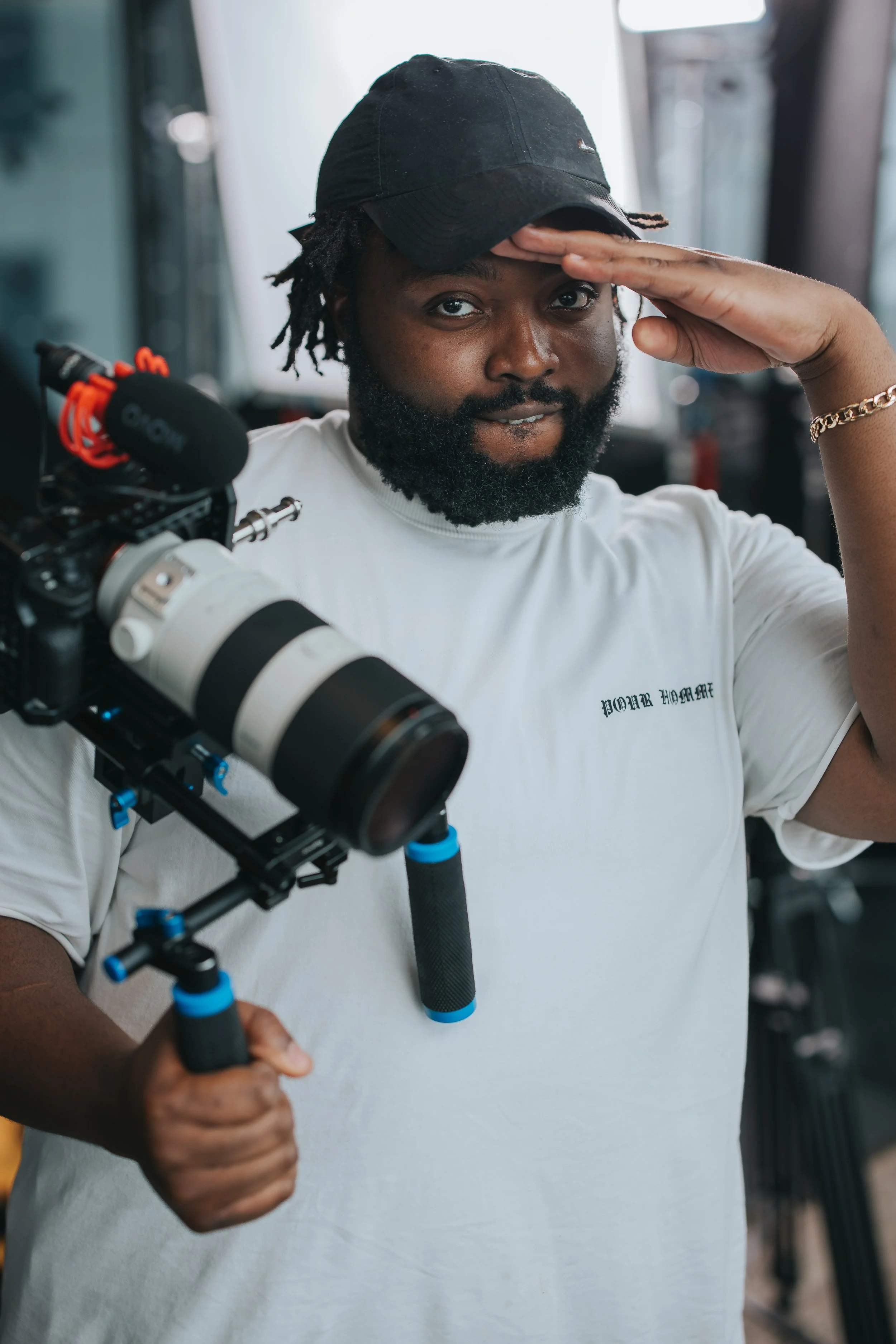 Man holding a professional camera with a large telephoto lens, saluting or shading his eyes with his hand, wearing a black cap and a white t-shirt.