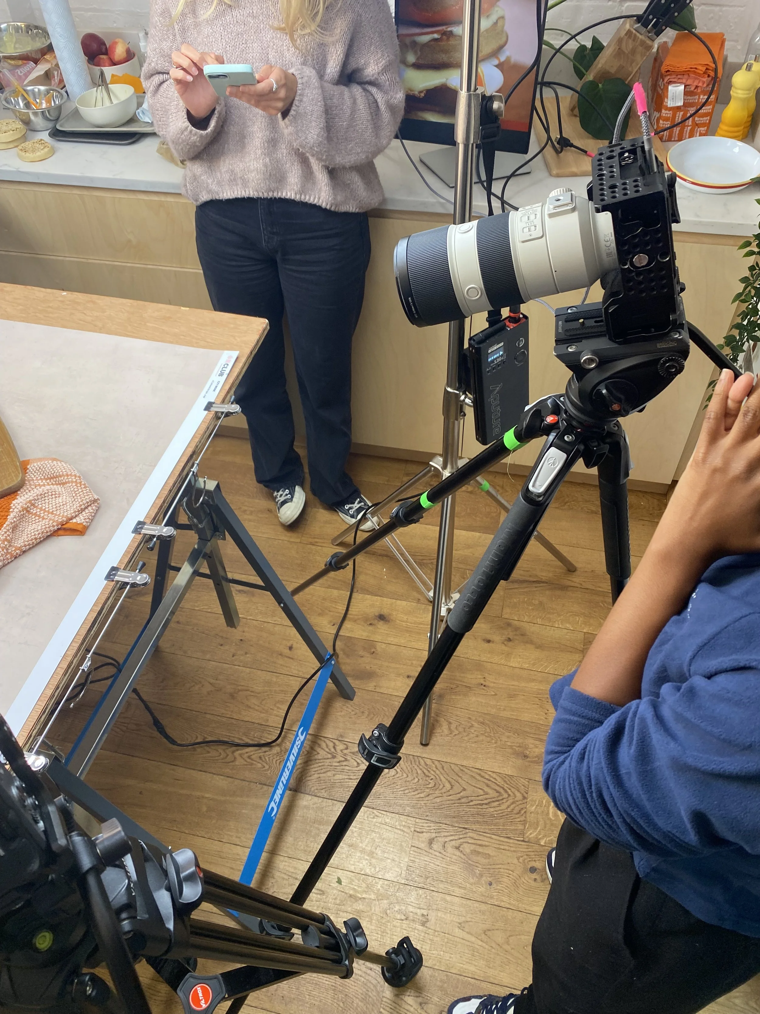 A person in a pink sweater and dark pants is holding a smartphone, standing in a kitchen with various food items and kitchenware on the counter. A camera on a tripod is set up nearby, indicating a photo or video shoot happening in the kitchen.