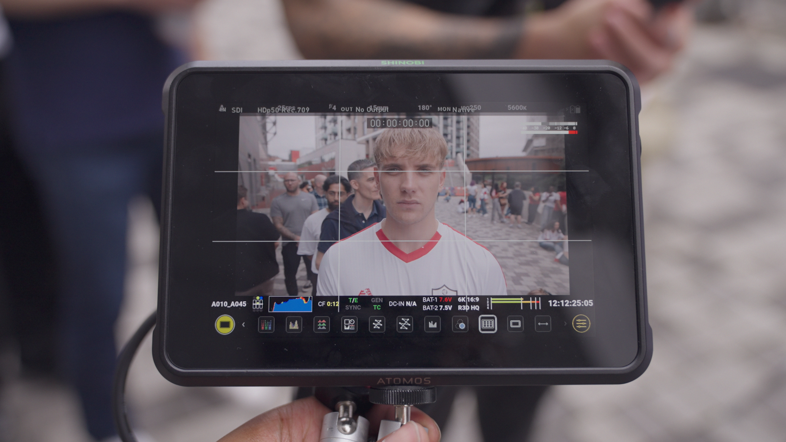 A camera monitor showing a young man with blonde hair and a red-and-white sports jersey, standing among a crowd outdoors in an urban setting.