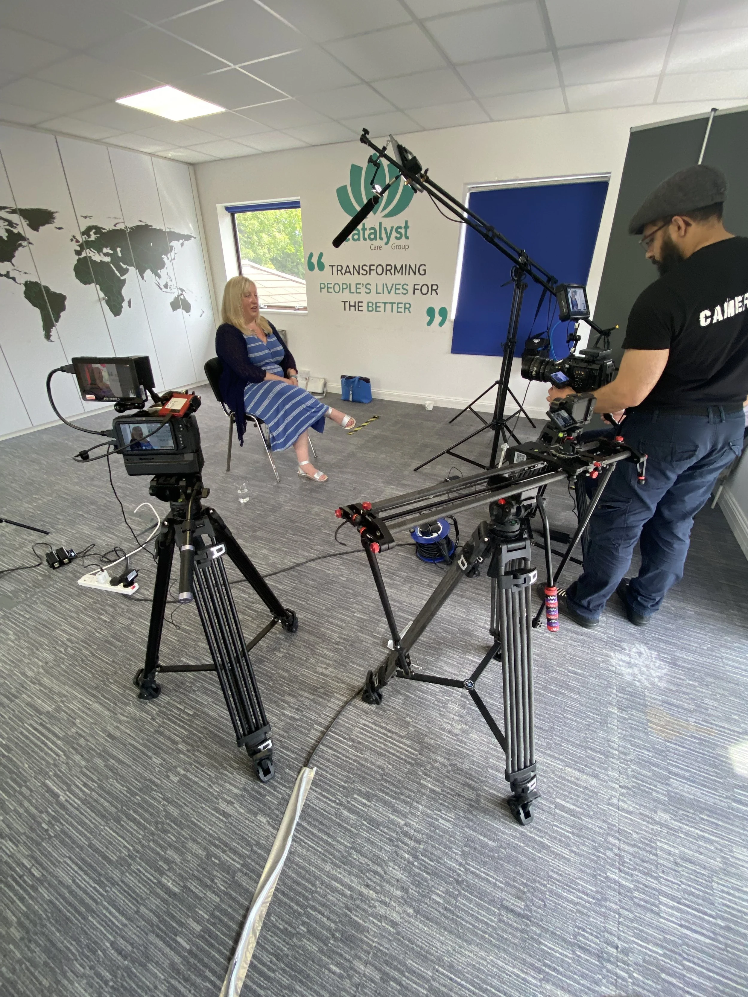 A woman is being filmed sitting on a chair in a room with a large world map on the wall and a sign that says 'Transforming People's Lives for the Better.' Two cameras and filming equipment are set up around her, and a crew member is operating one of 