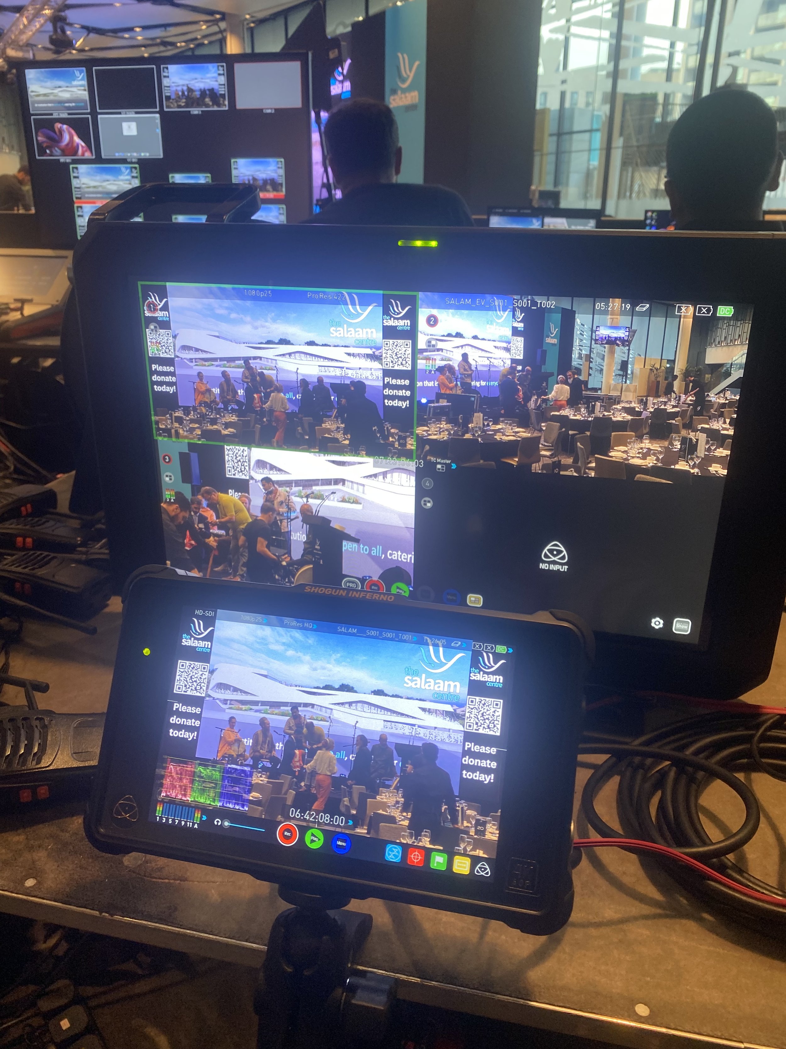 A video production setup at an event with multiple monitors and a tablet capturing the scene. The scene shows a conference or banquet with people seated at tables, a stage with a large display, and branding for Salaam