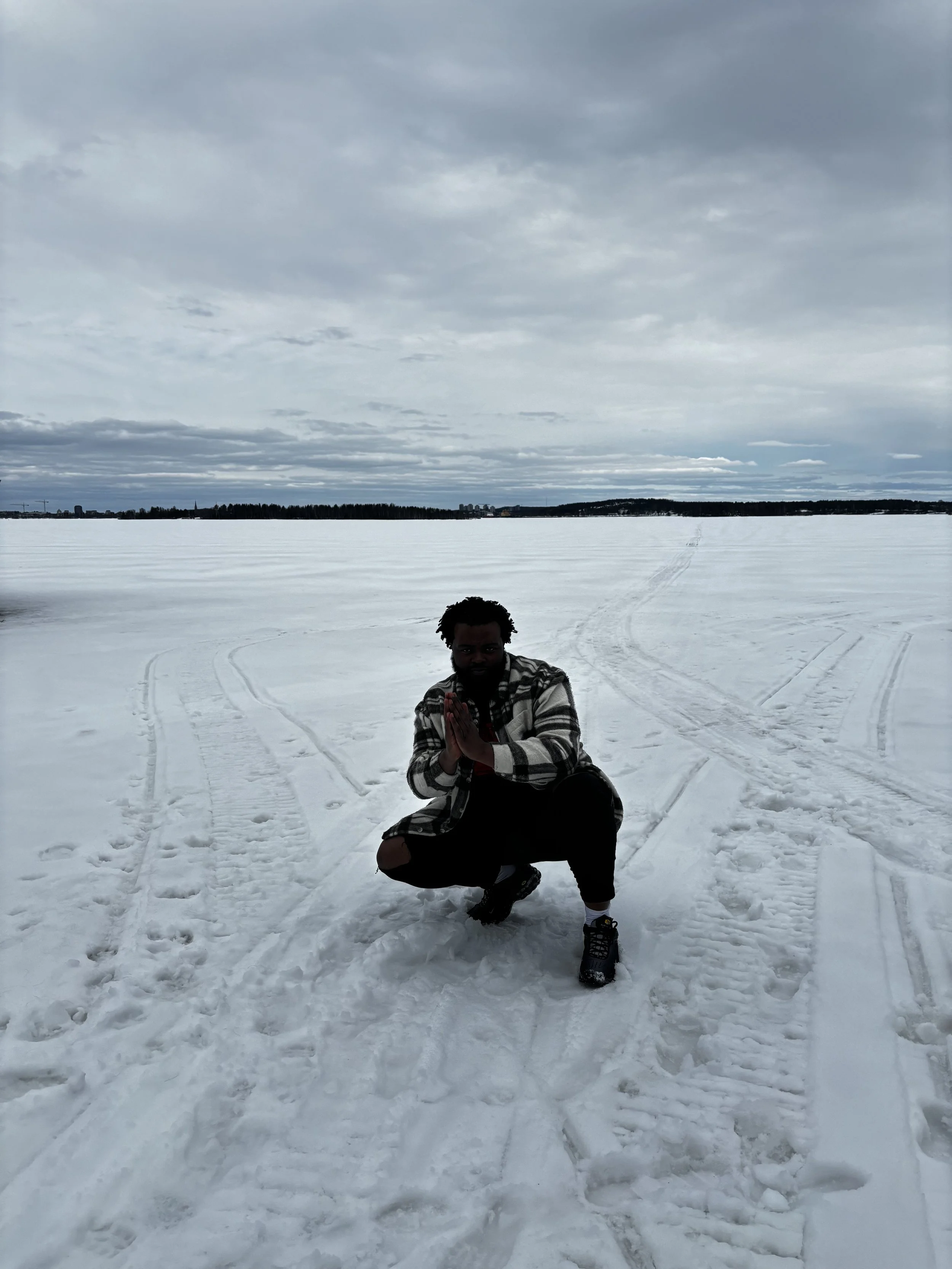 Person crouching on snowy ground with a snow-covered landscape and cloudy sky in the background.
