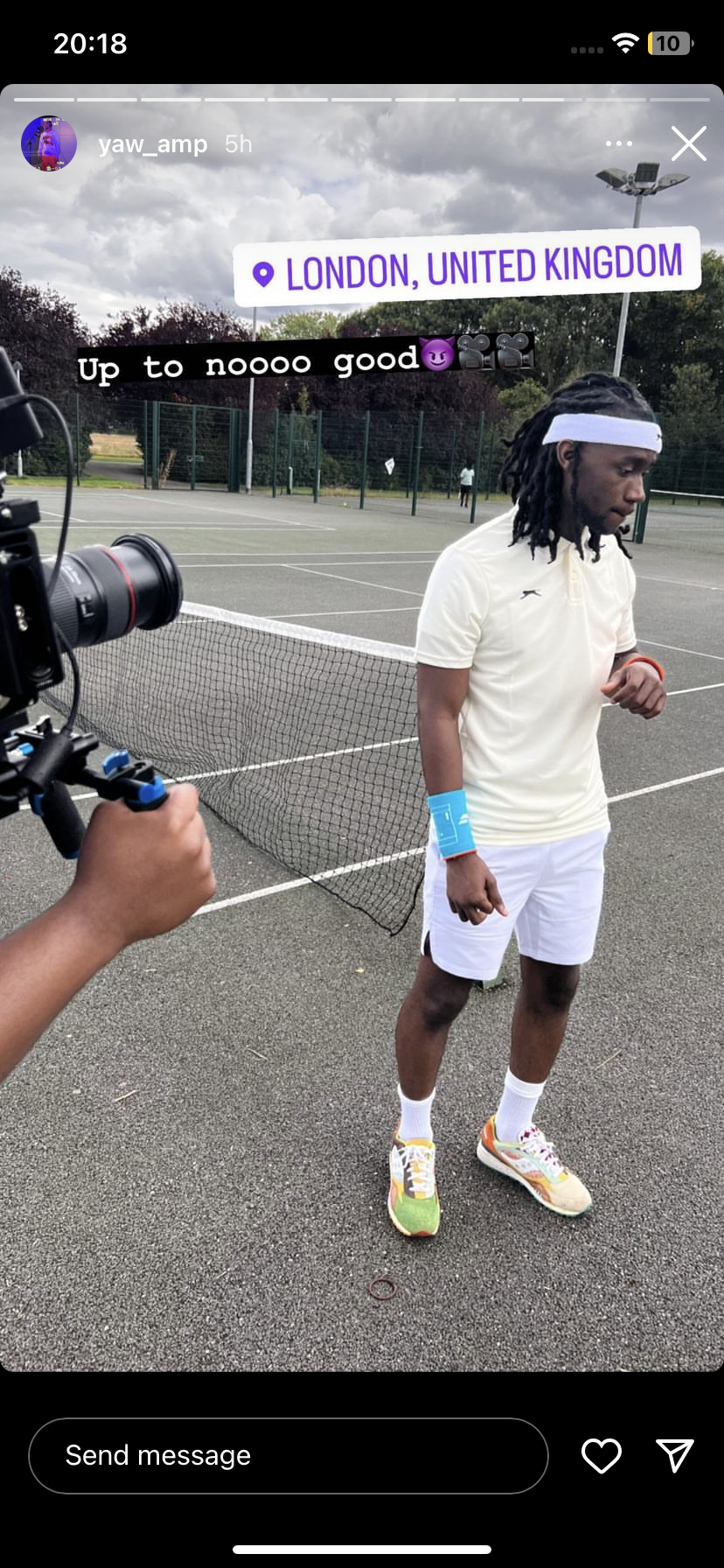 A man on a tennis court wearing a white sports outfit with colorful sneakers, standing near the net, with a camera person capturing him. The location tag shows London, United Kingdom. The sky is cloudy.