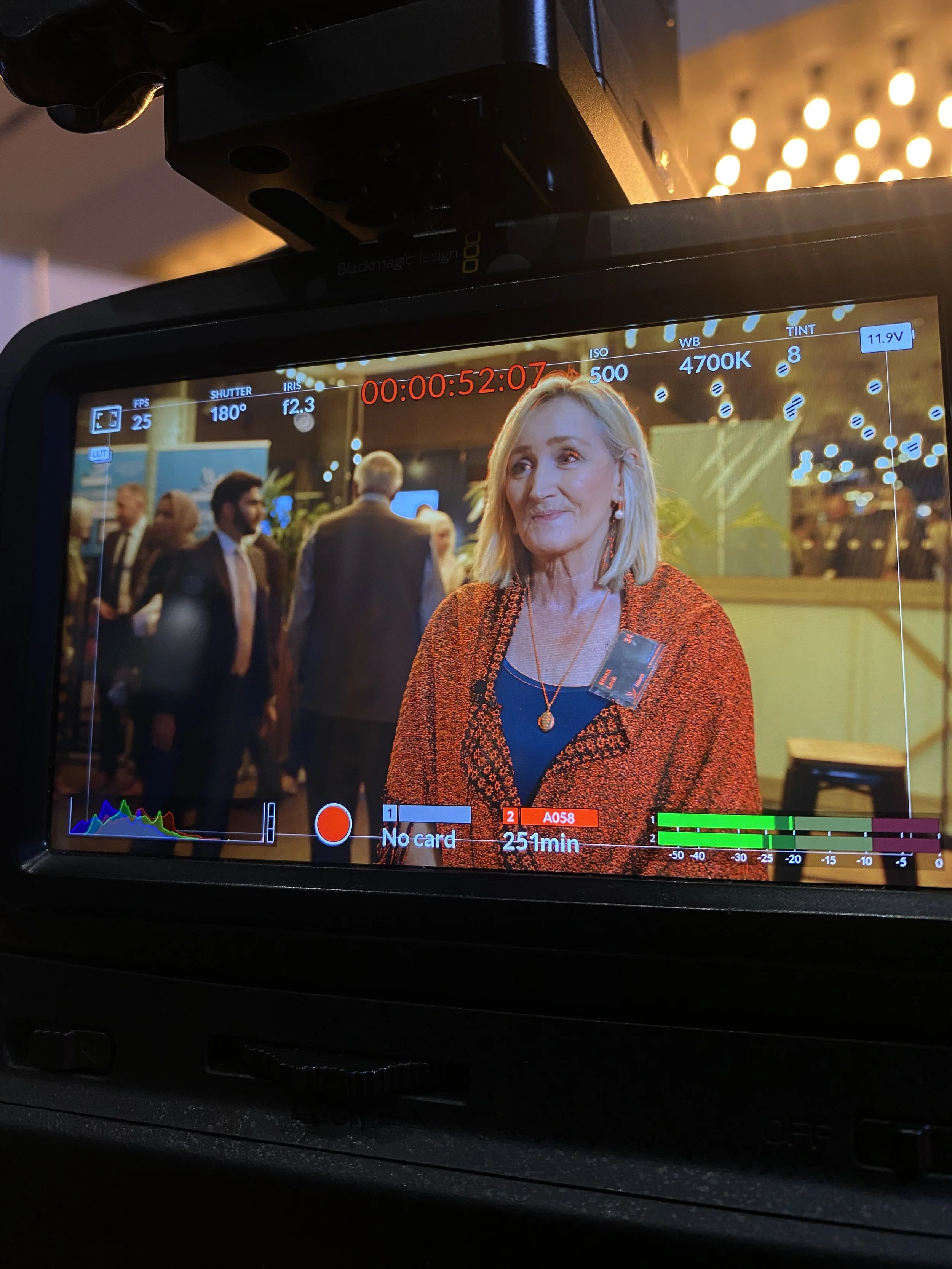 A woman standing at an event, being recorded by a video camera, with people in suits in the background.