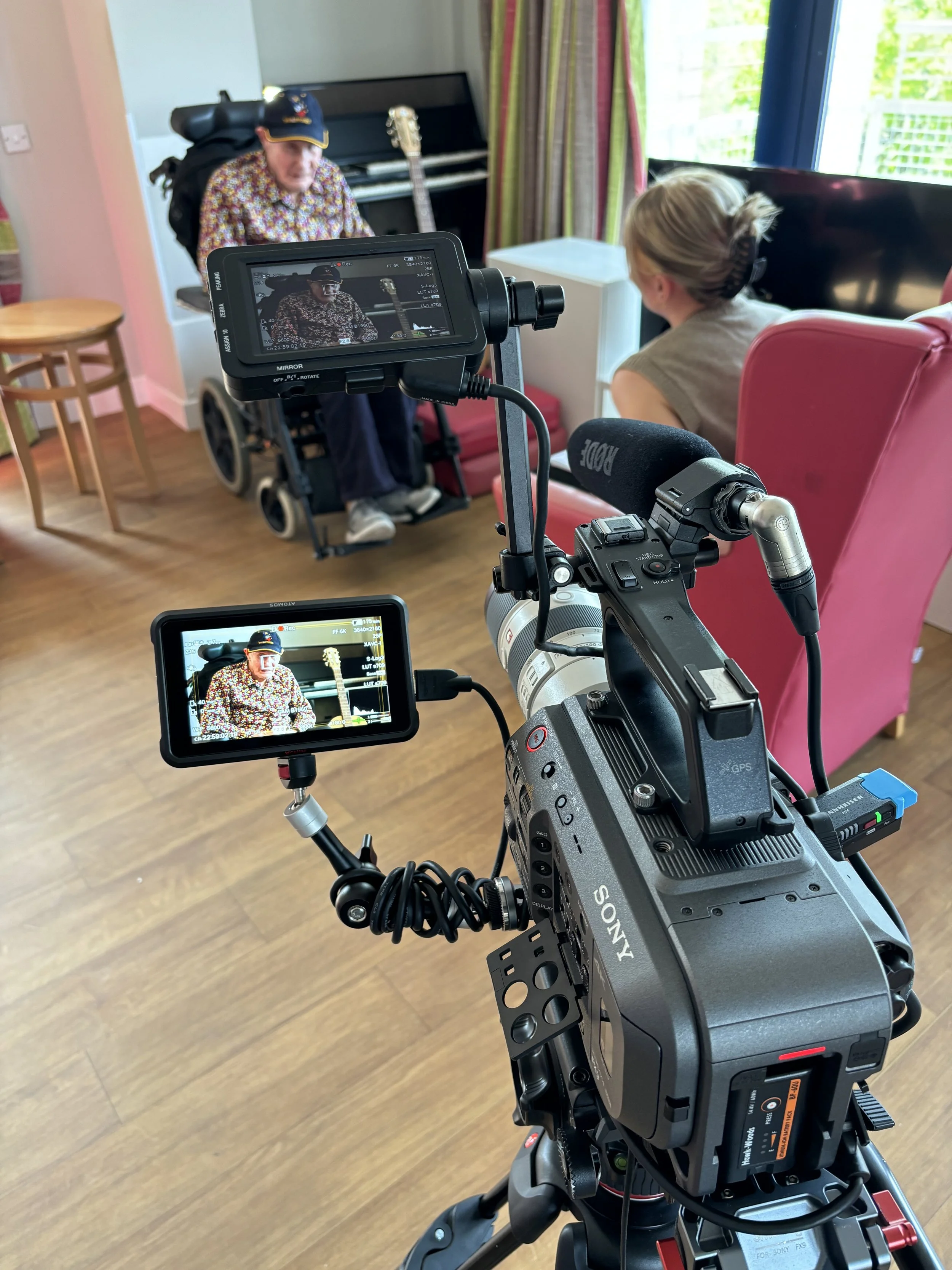 A video camera recording a man in a wheelchair wearing a floral shirt and a cap, with a woman sitting nearby in a pink armchair, in a room with a piano, curtain, and window with blinds.