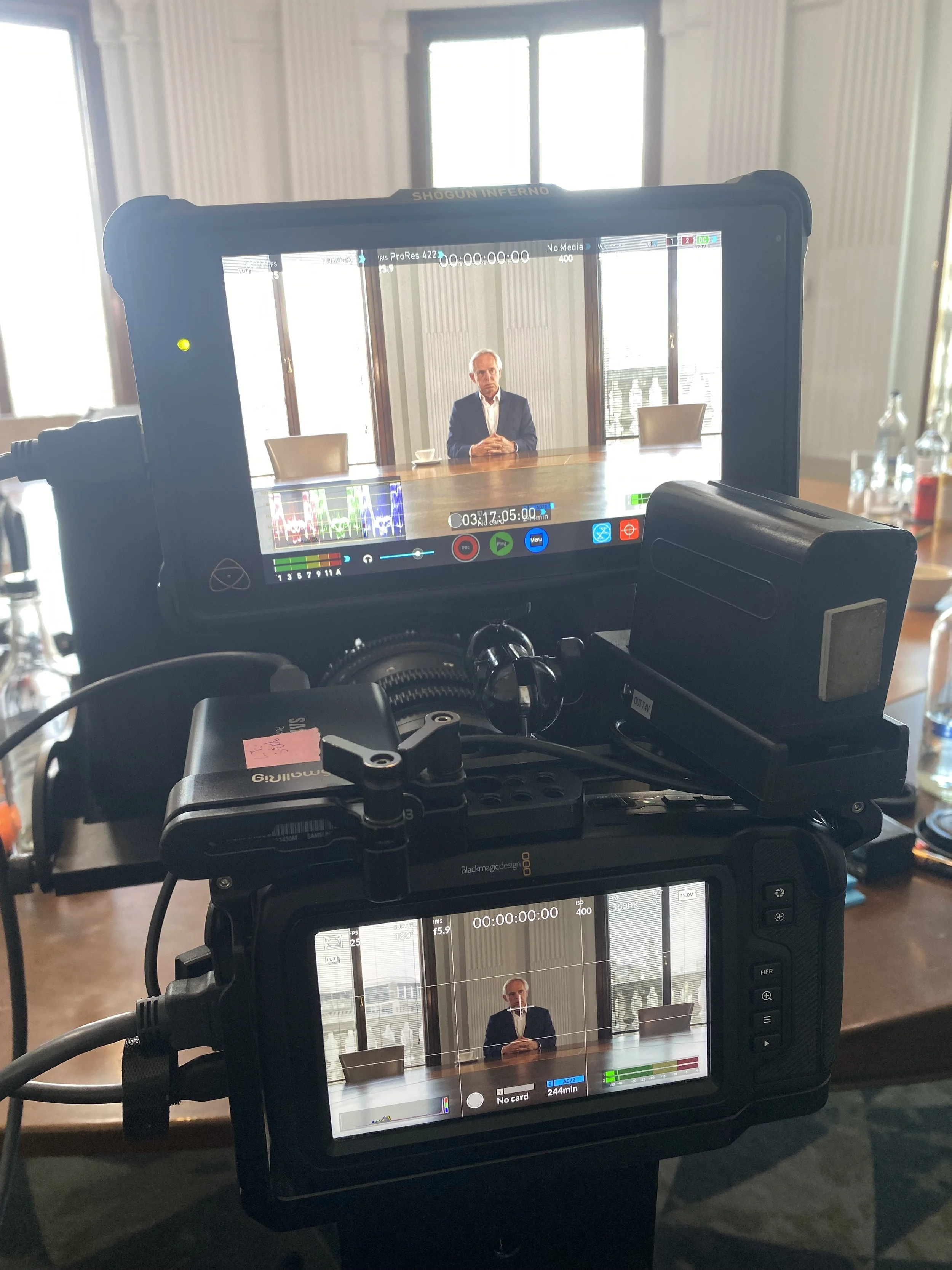 Two cameras focused on a man in a suit sitting at a conference table with a window and balcony in the background.
