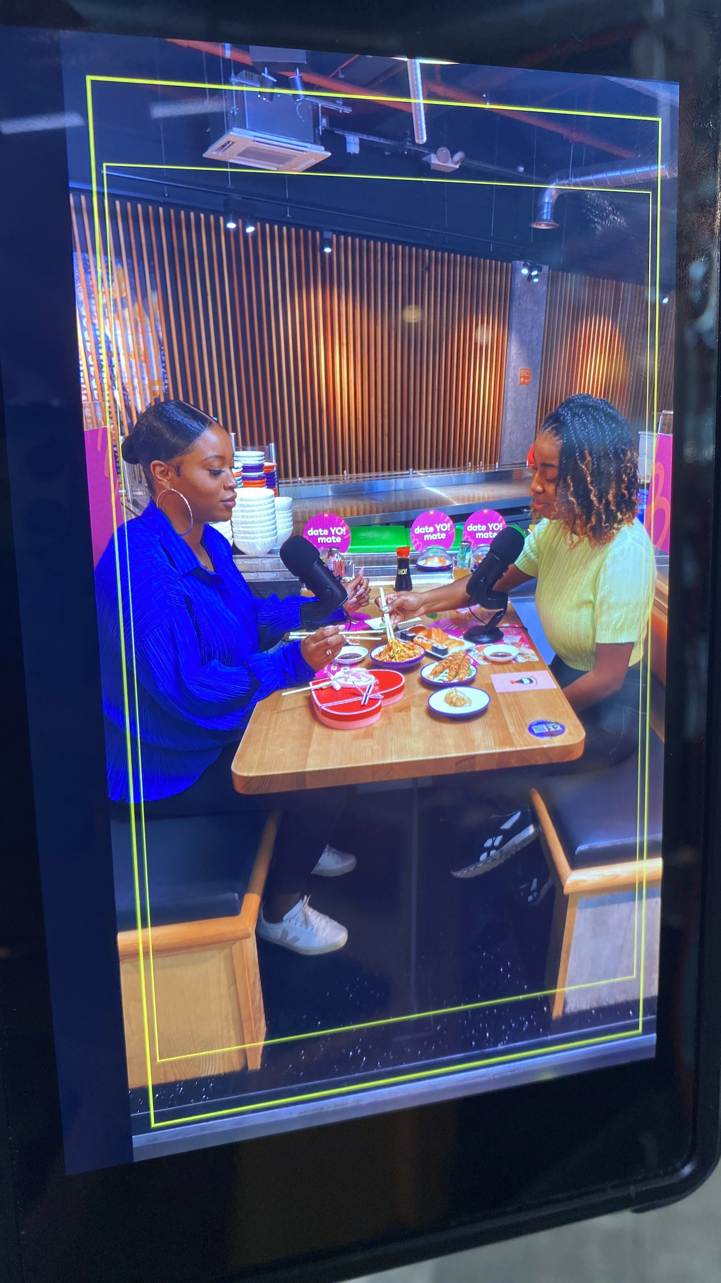 Two women having sushi at a restaurant table with microphones, plates, and drinks, viewed through a glass screen.