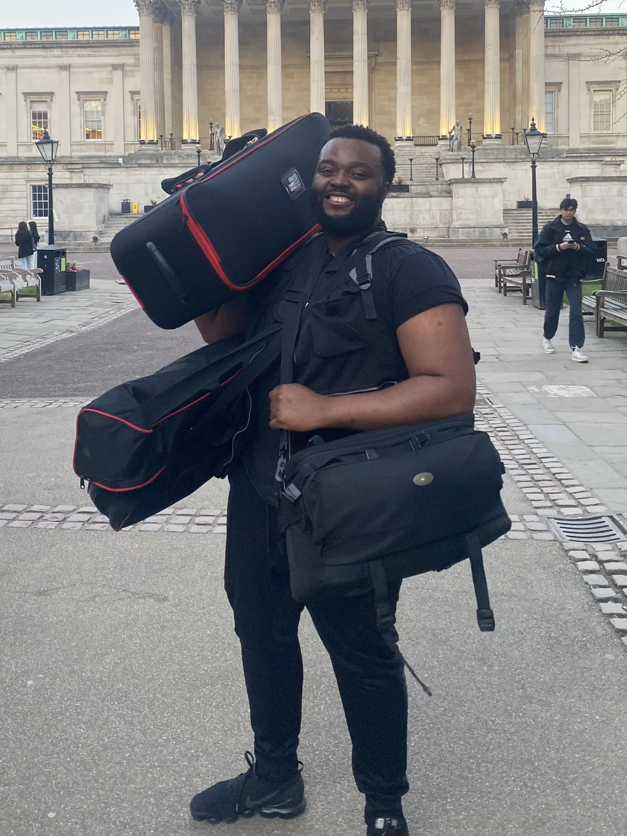A smiling man carrying a large black and red suitcase on his shoulder and holding a black bag in his hand, standing outside in front of a classical building with columns.