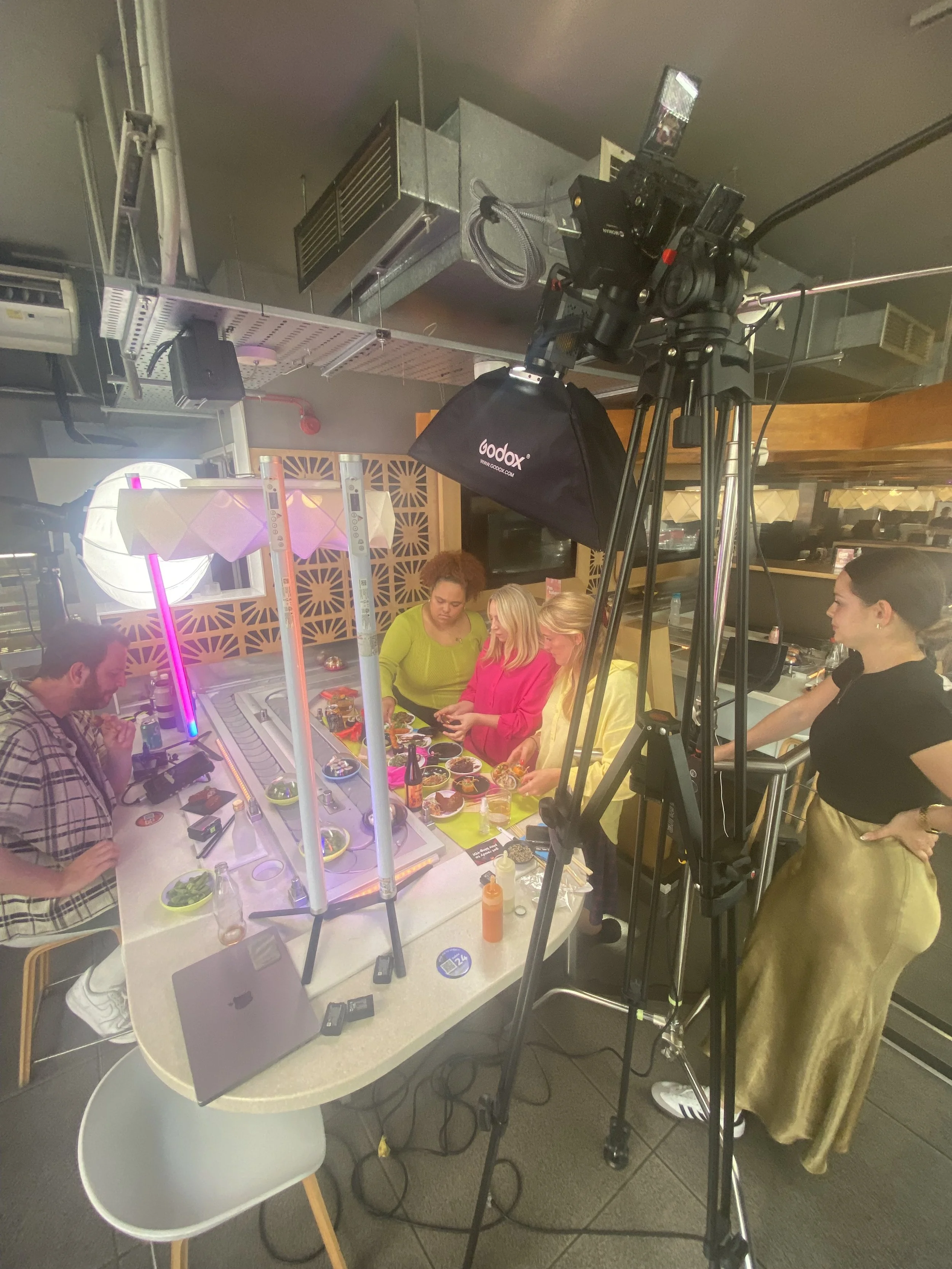 A behind-the-scenes view of a camera and lighting setup during a live recording or photo session in a restaurant or cafe. Several women are gathered around a table with food and drinks, and a woman on the right appears to be directing or managing the