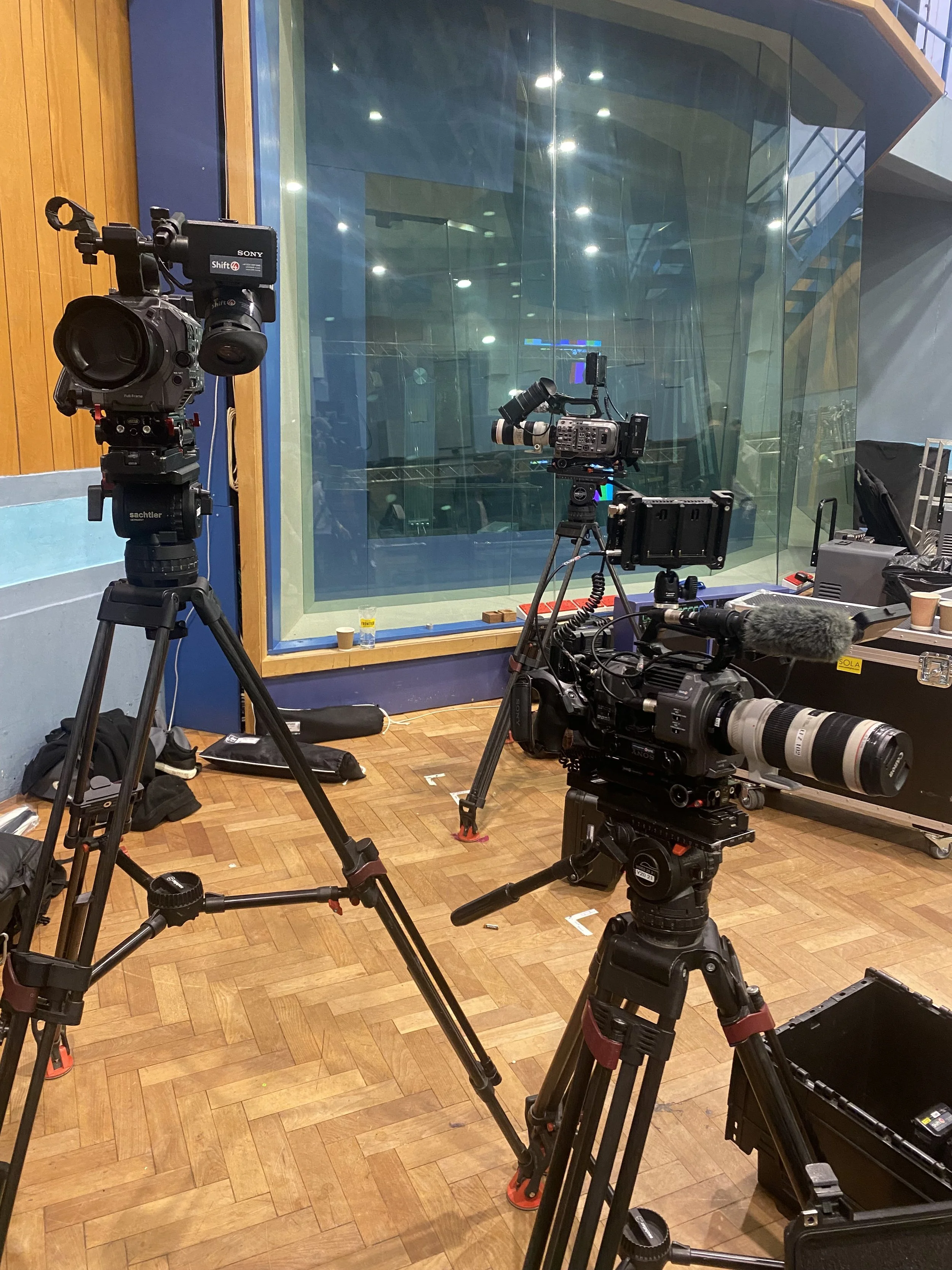 Two professional video cameras on tripods set up in a recording studio with a large glass window showing an empty room with wooden floors, some equipment, and a staircase.