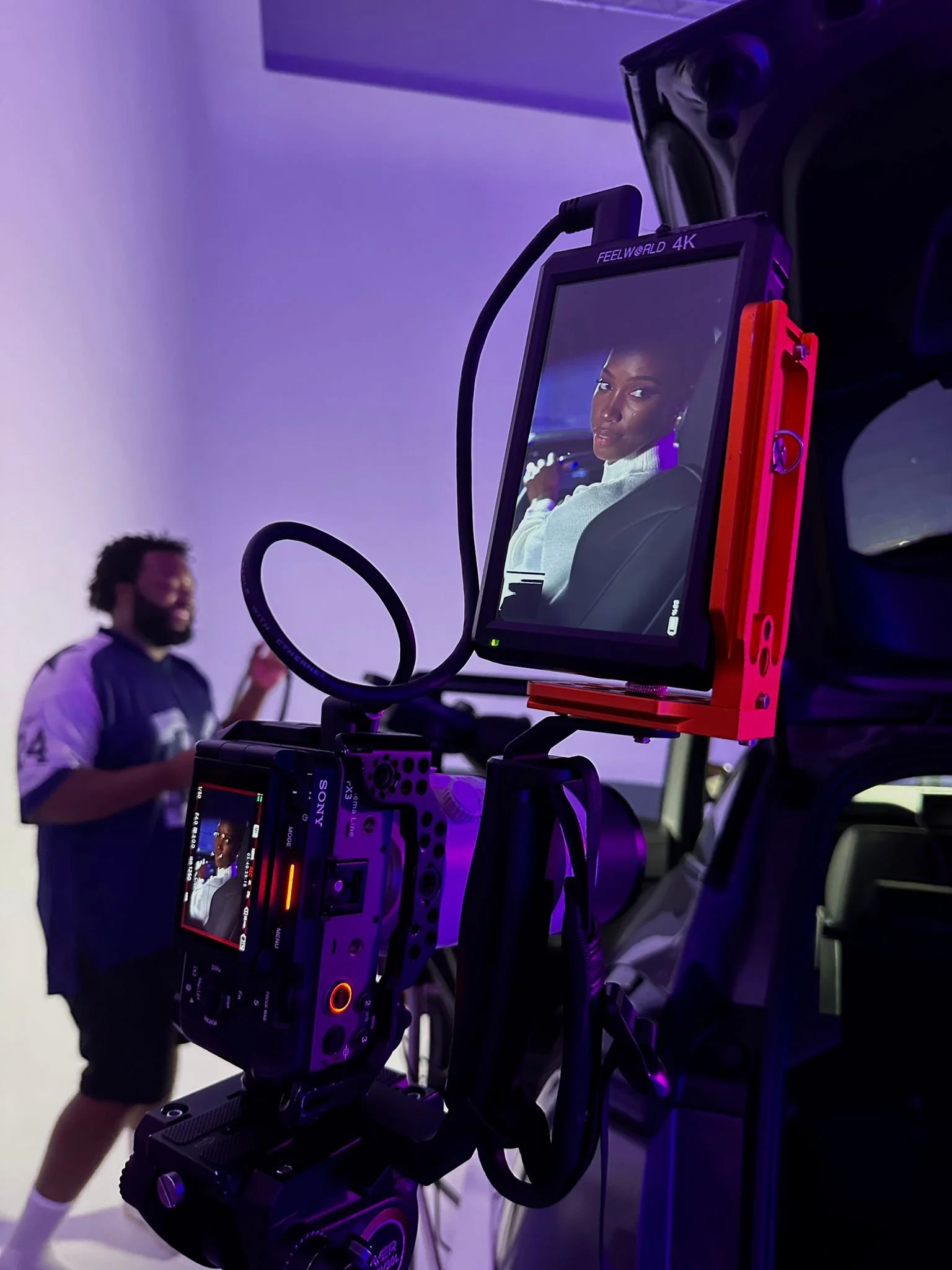 A professional video camera capturing a woman sitting in a car, with a man in the background on a set with purple lighting.