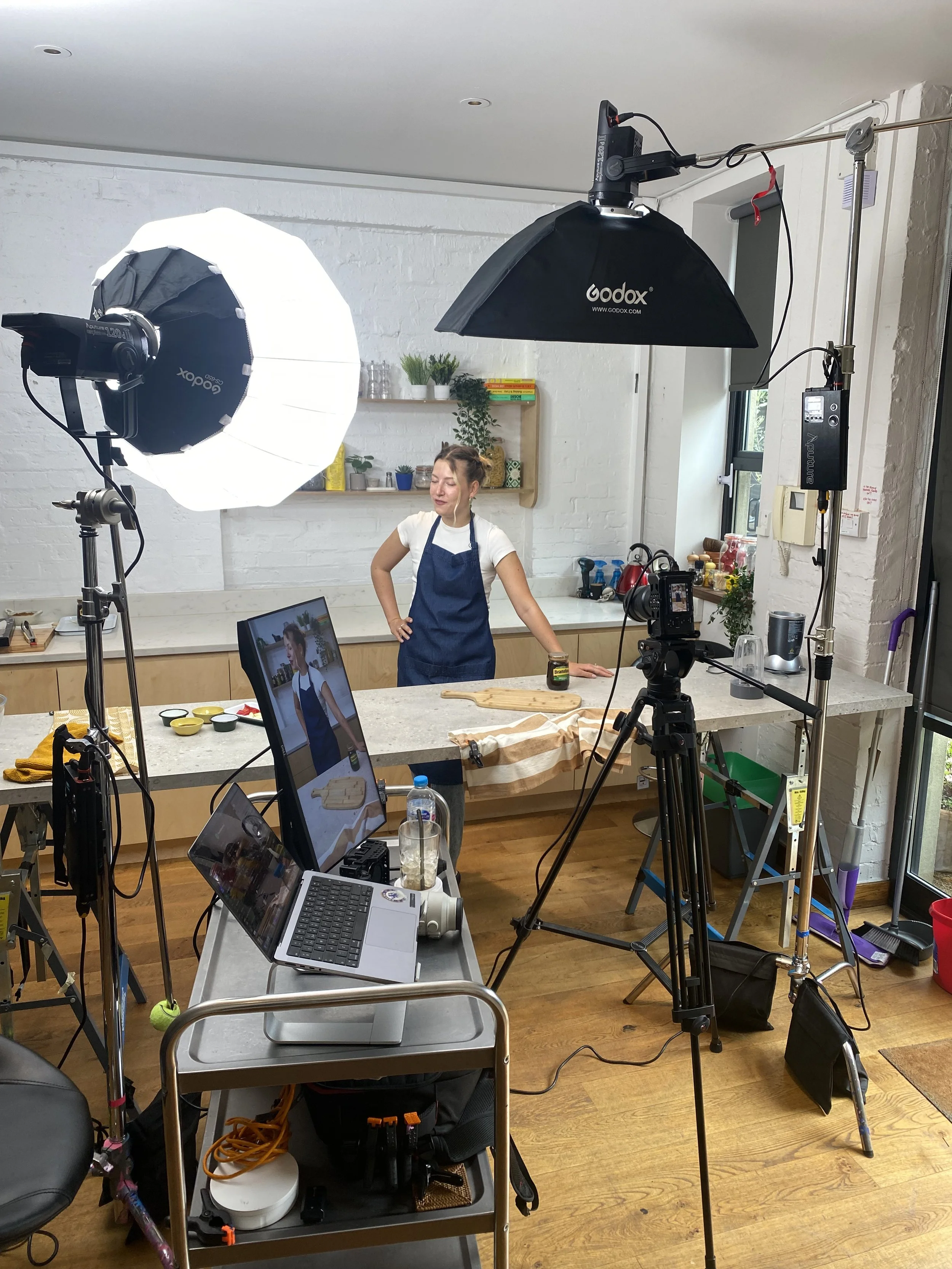 A woman wearing a blue apron stands in a well-lit kitchen while being filmed with professional lighting and camera equipment. The setup includes large softbox lights, a camera on a tripod, and a laptop on a rolling cart. The white brick wall in the b