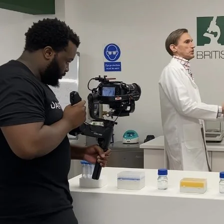 A man filming with a professional camera in a laboratory, observing a scientist who is working at a lab bench with test tubes and lab equipment.