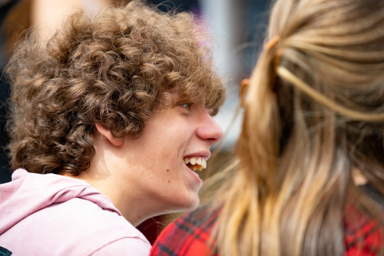 Profile of a boy with light brown curly hair smiling and the back of a blondes head in the corner