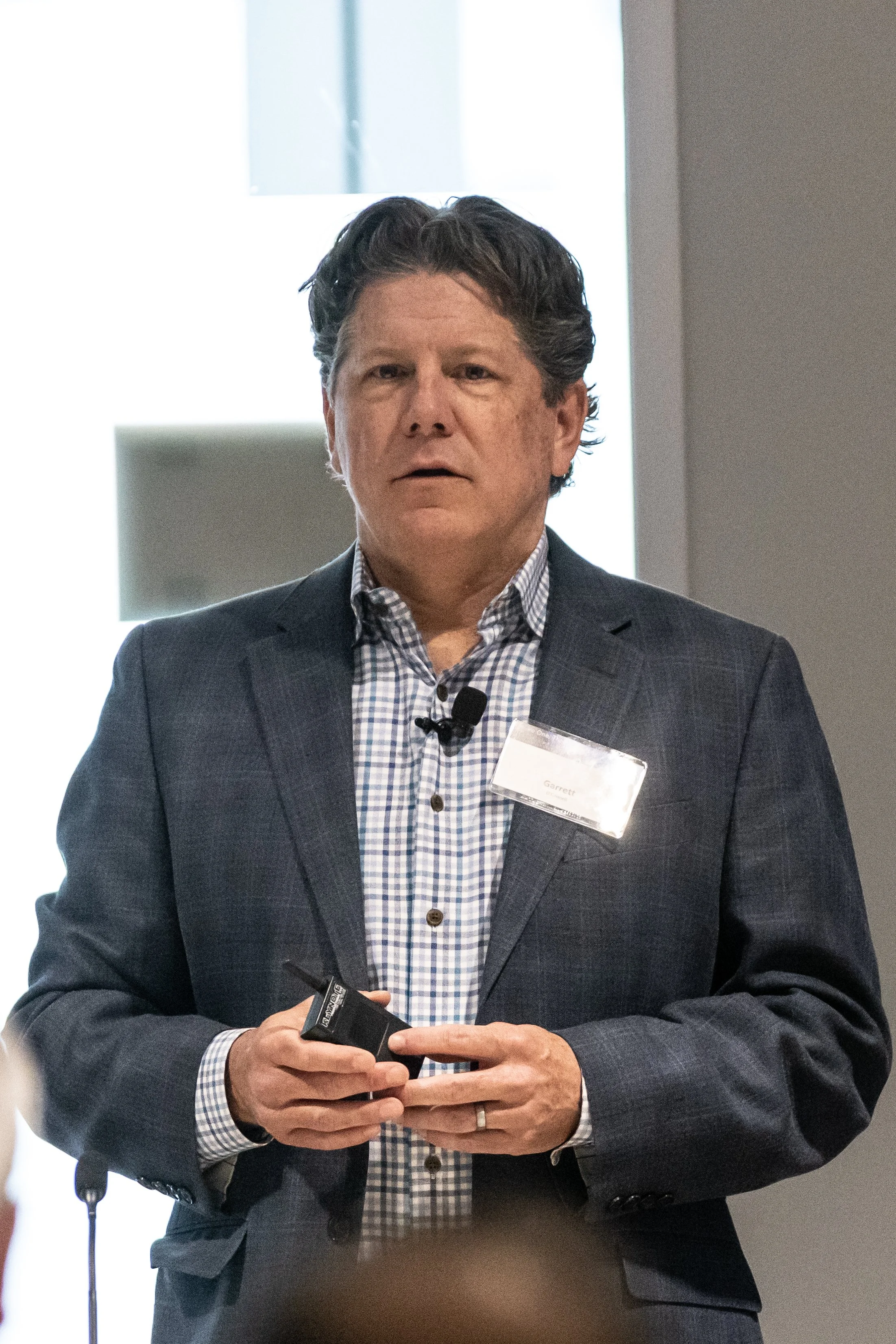 A man in a dark suit and checkered shirt standing in a well-lit indoor setting, holding a small black device in his hands, presenting at an event.