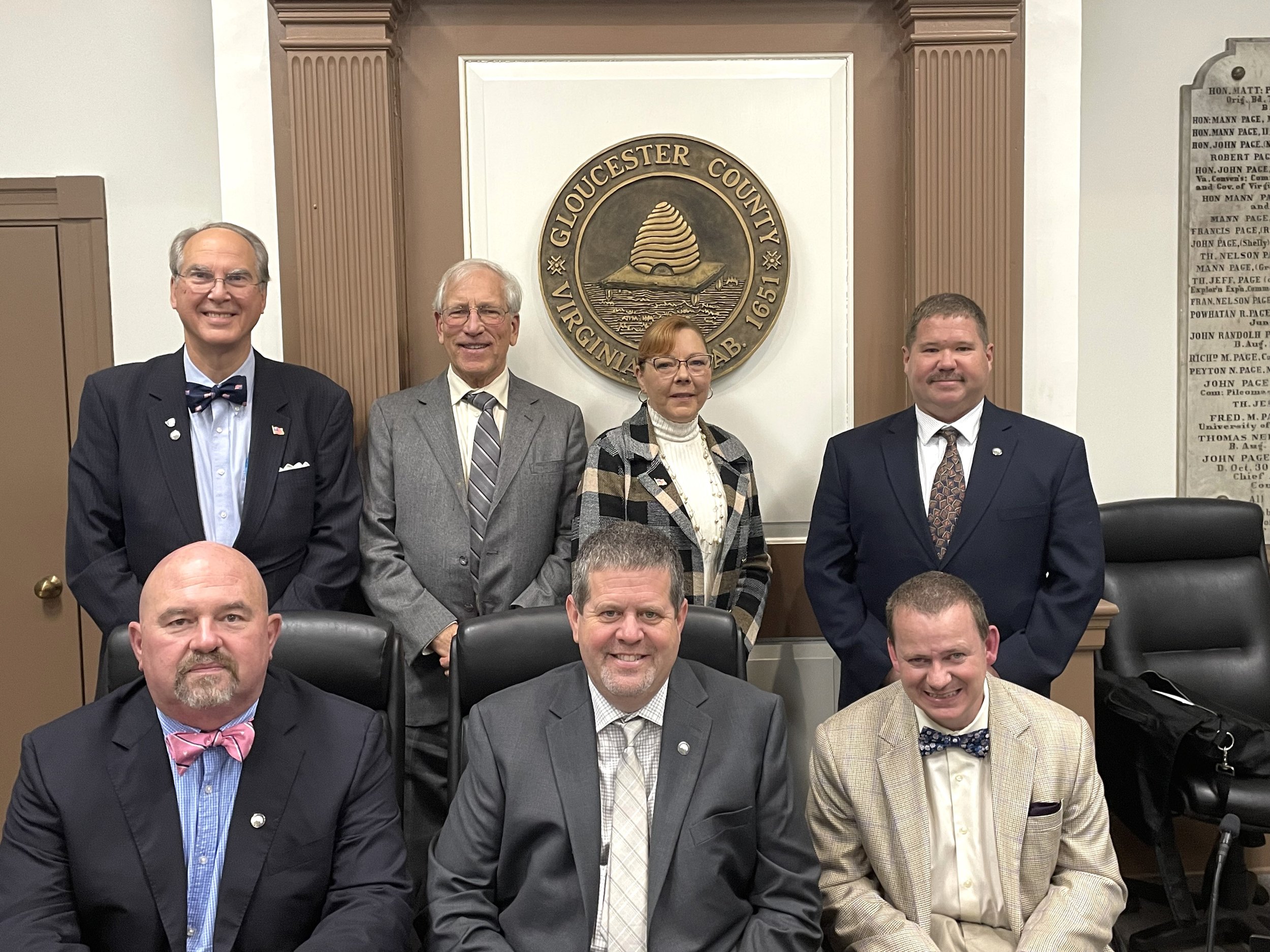Six men and one woman in front of a County seal