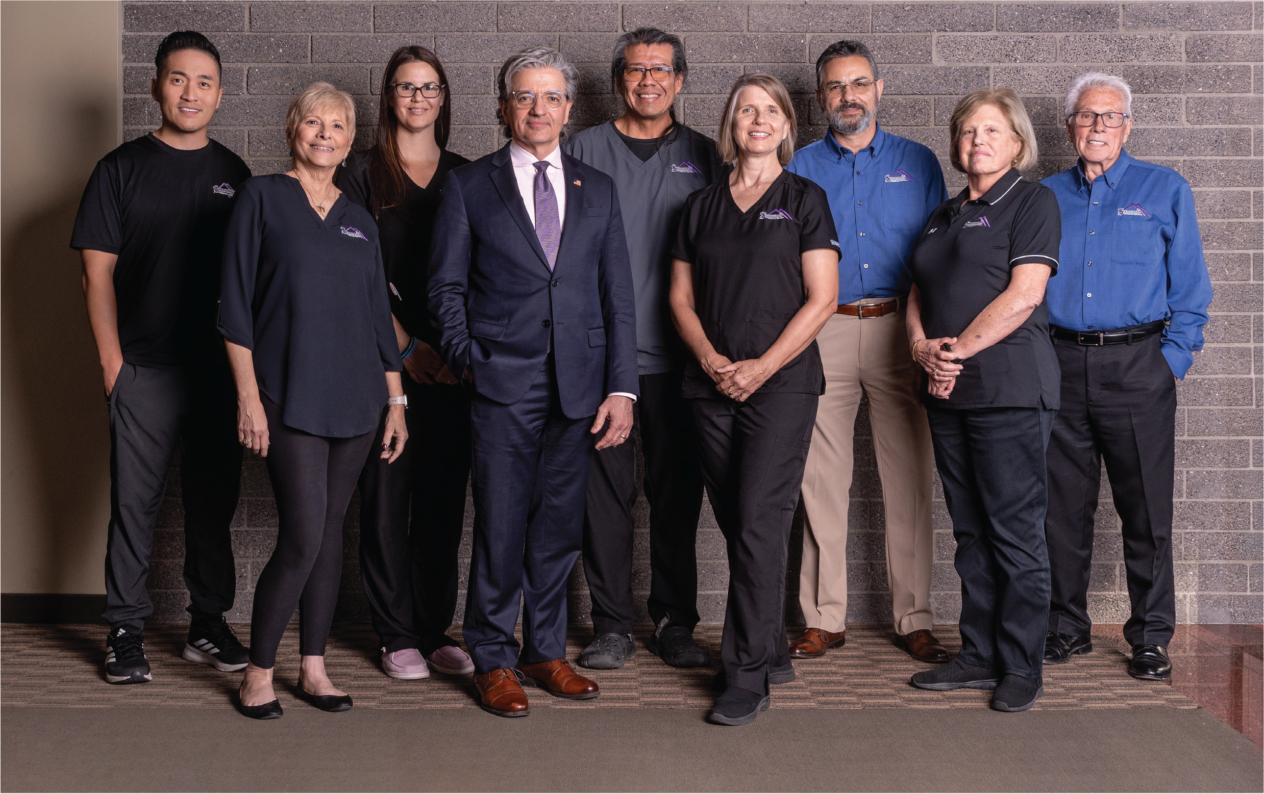 A diverse group of ten people standing against a brick wall, smiling for the camera. The group includes men and women of varying ages, dressed in casual and business attire.