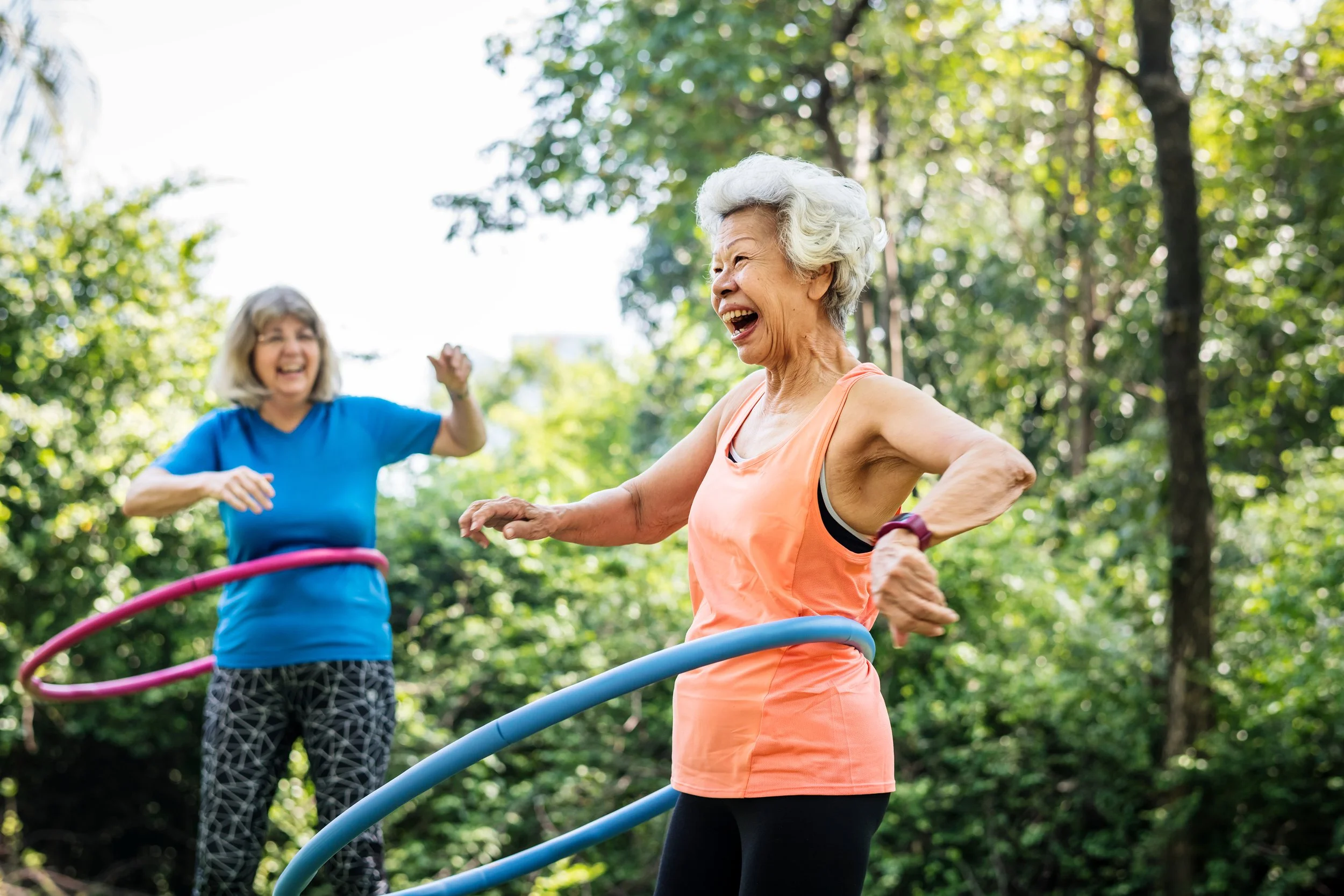 Two elderly women having fun playing with hula hoops outdoors in a green park, smiling and laughing.