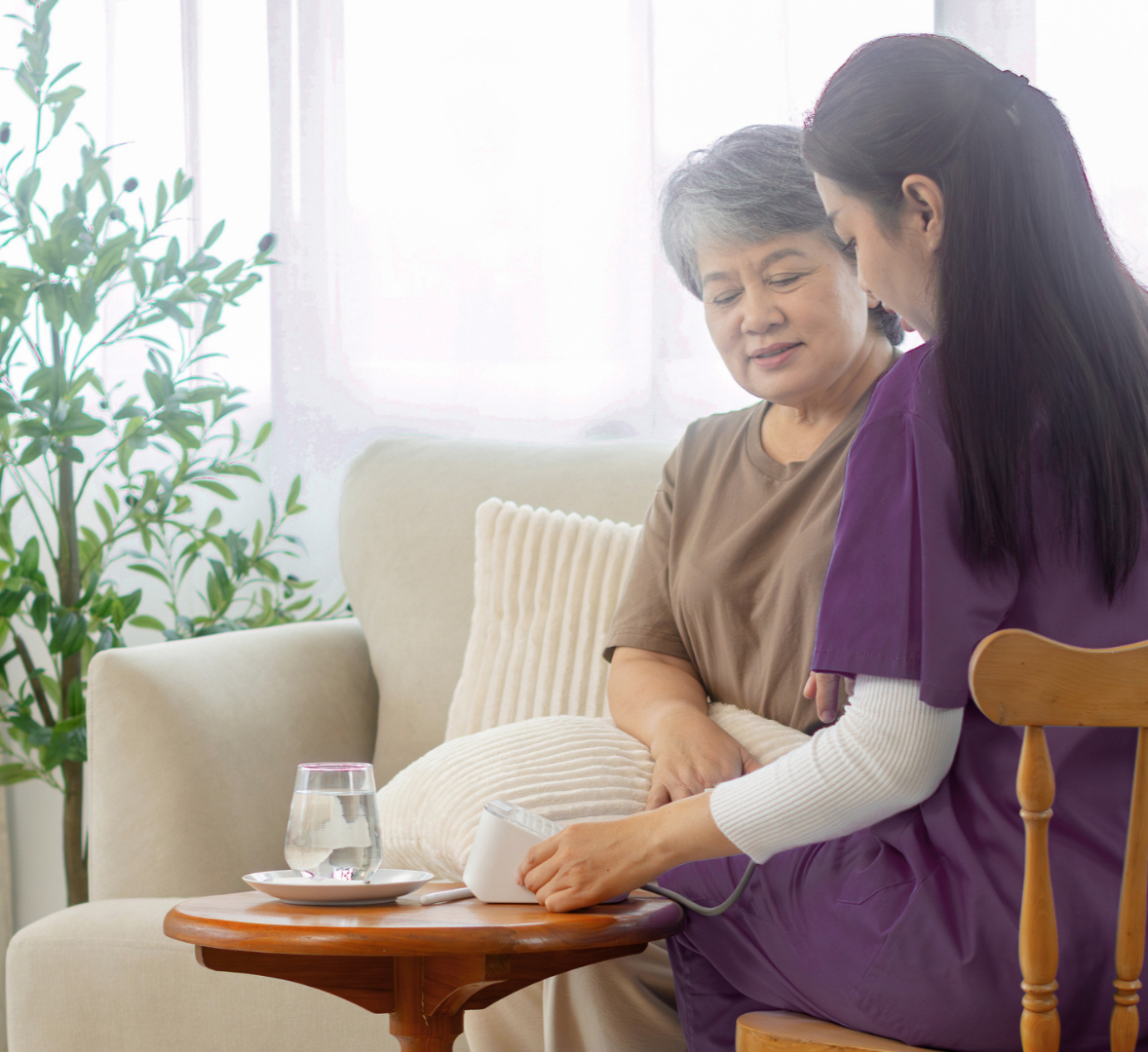 A caregiver using a blood pressure monitor on an elderly woman sitting on a sofa in a bright living room.