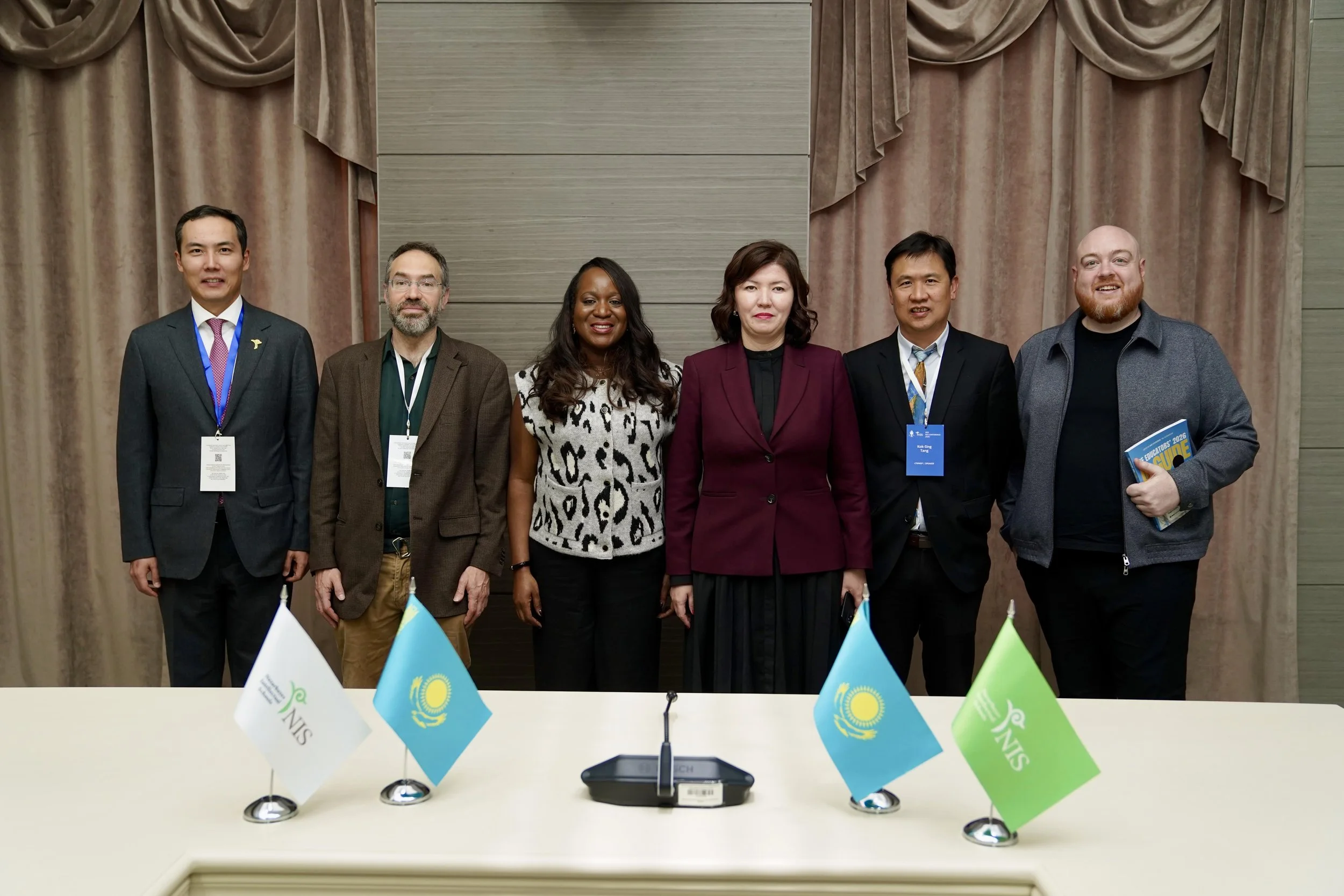 Toju Duke and AI professors from Australia, and the Minister of Education Kazakhstan - Zhuldyz Suleimenova standing behind a conference table with flags of Kazakhstan displayed in front of them, in a conference room with beige curtains. 