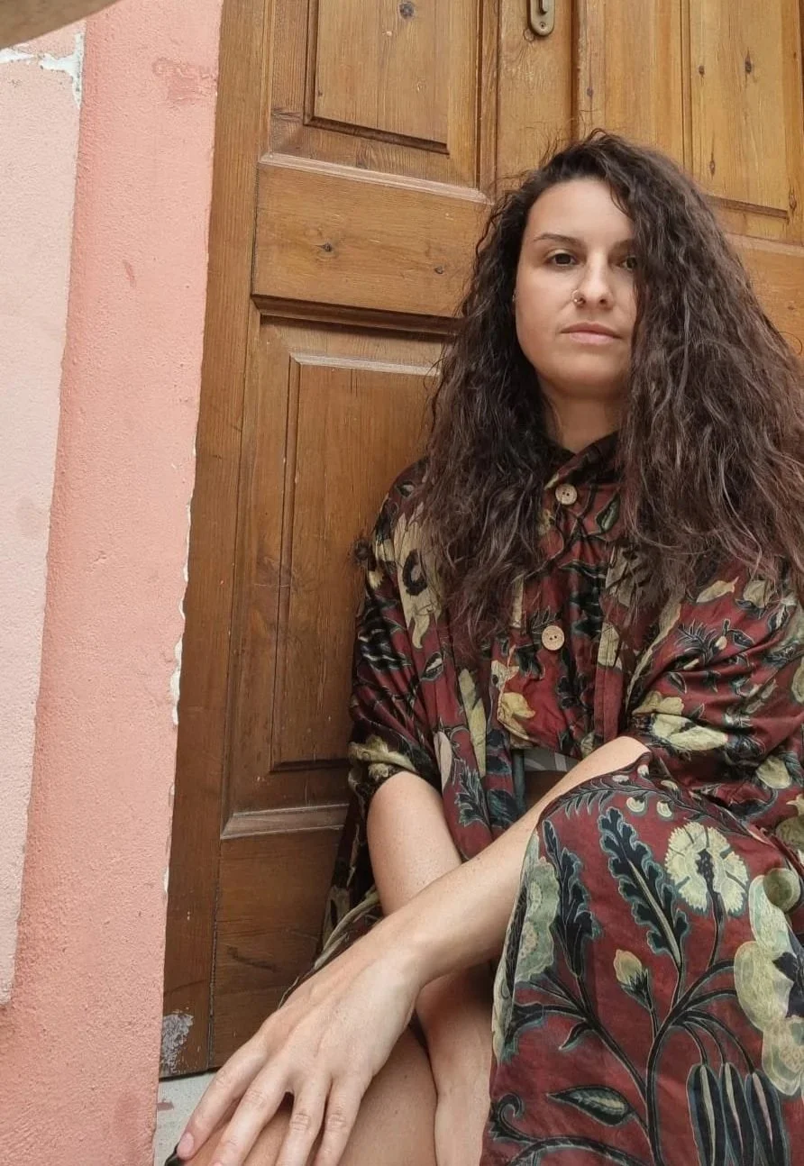 Una donna con i capelli ricci scuri e indossa un abito con motivi floreali seduta accanto a una porta di legno.