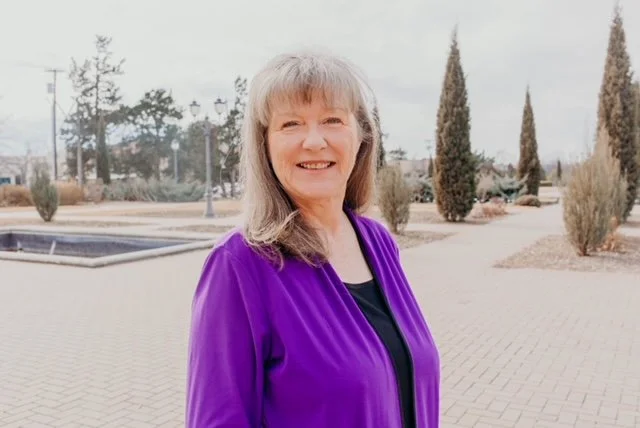 A smiling woman with gray hair wearing a purple jacket standing outdoors in a park-like setting with sparse trees and bushes.