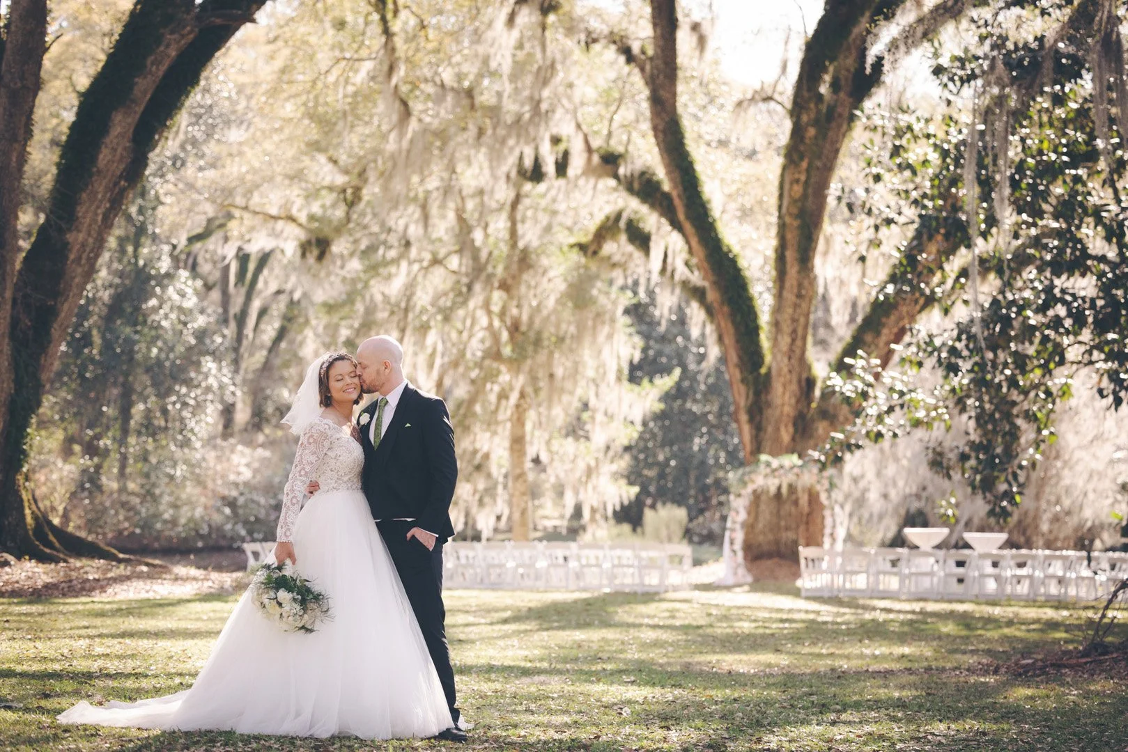 Bride and groom standing together in a garden, with large trees in the background, she wears a lace wedding dress and holds a bouquet, while he wears a black suit.