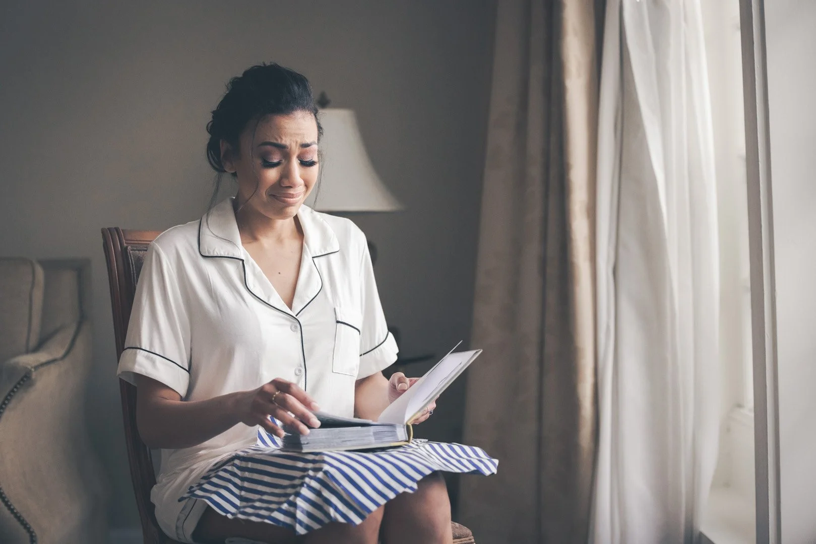Woman sitting indoors in pajamas, reading a book, and showing emotion.