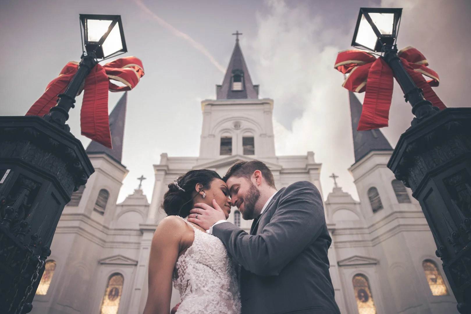 Bride and groom embracing in front of a historic church with red ribbons on lampposts.