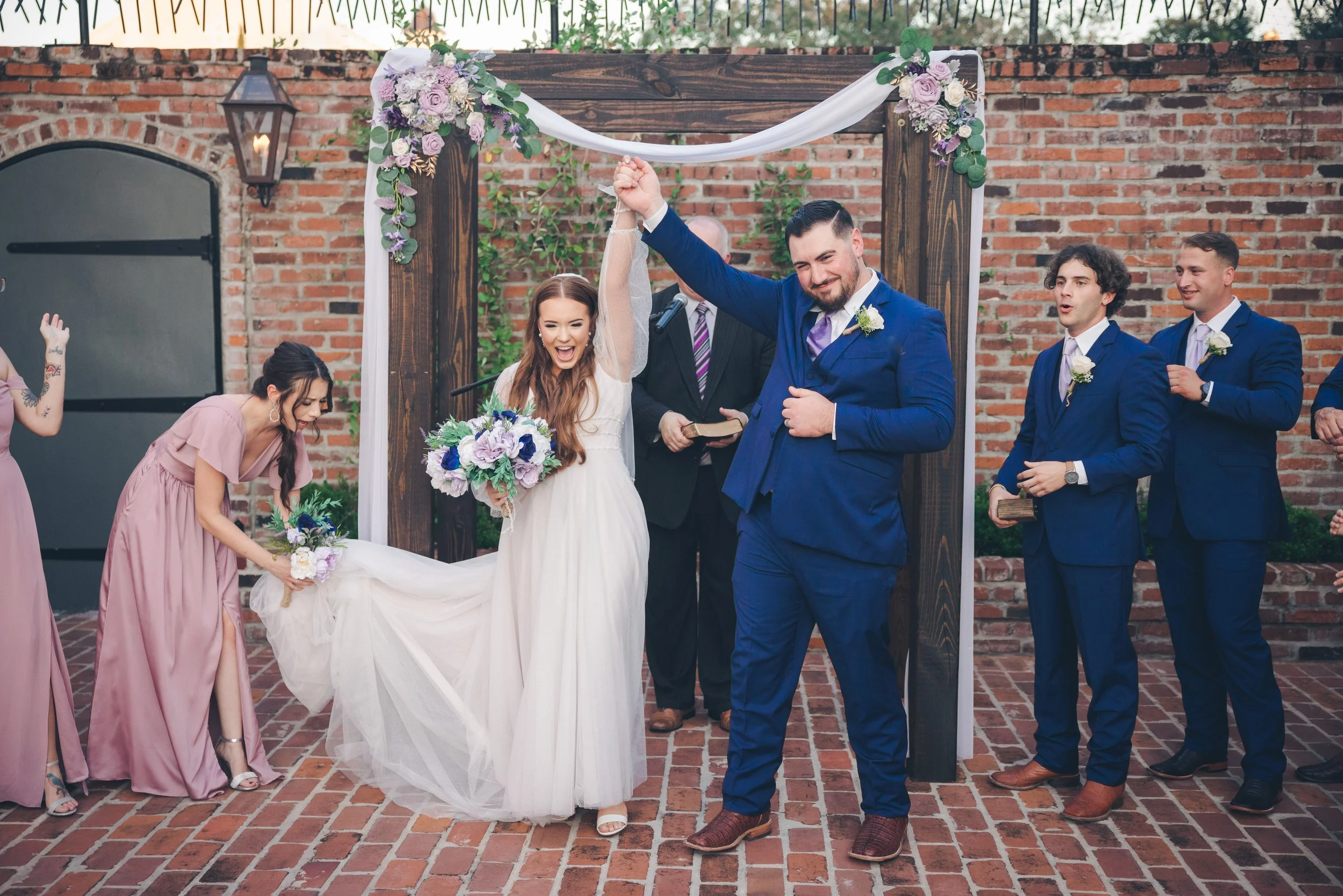 A joyful wedding scene with a bride in a white gown holding a bouquet and a groom in a blue suit, raising their hands in celebration under a floral arch, surrounded by bridesmaids in pink dresses and groomsmen in blue suits on a brick patio.
