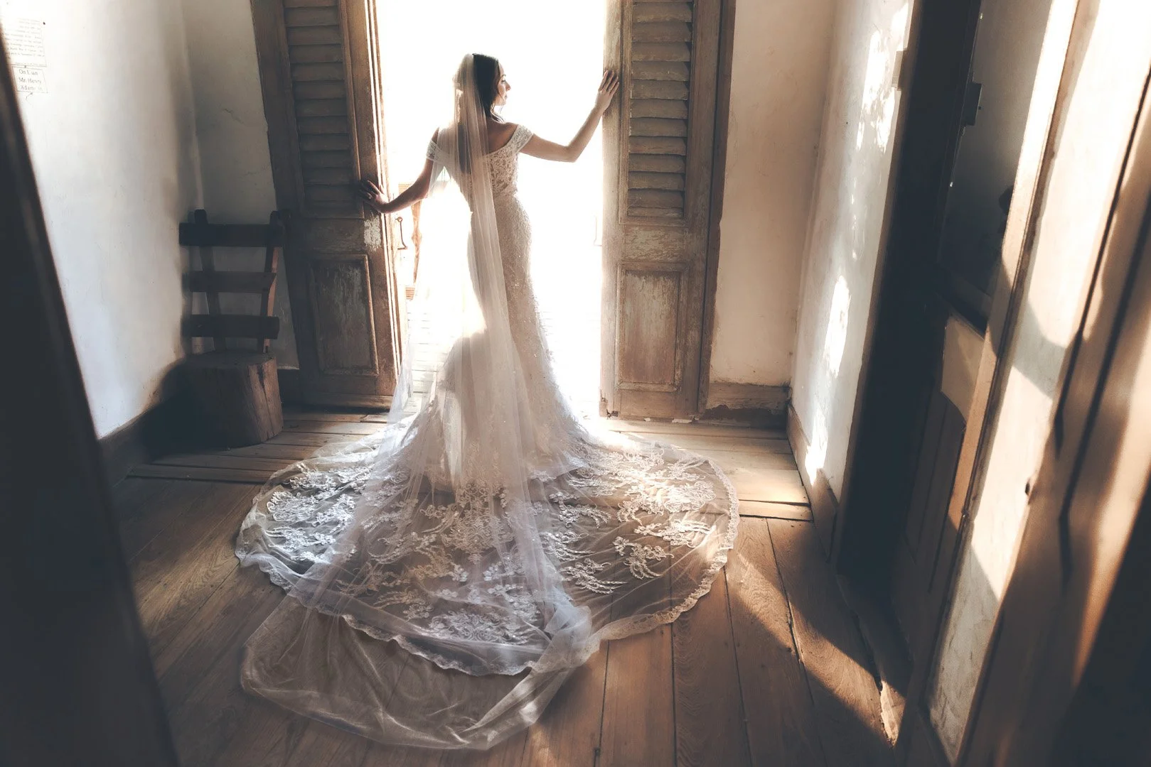 Bride in wedding dress standing in doorway with lace train, vintage wooden interior.