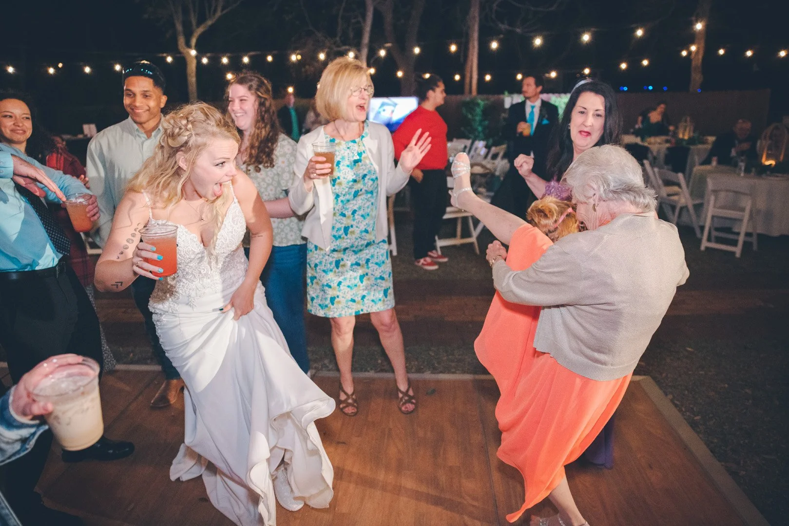 A group of people dancing at a wedding reception outdoors at night. A bride in a white dress is laughing while holding a drink, surrounded by guests with beverages, under string lights.