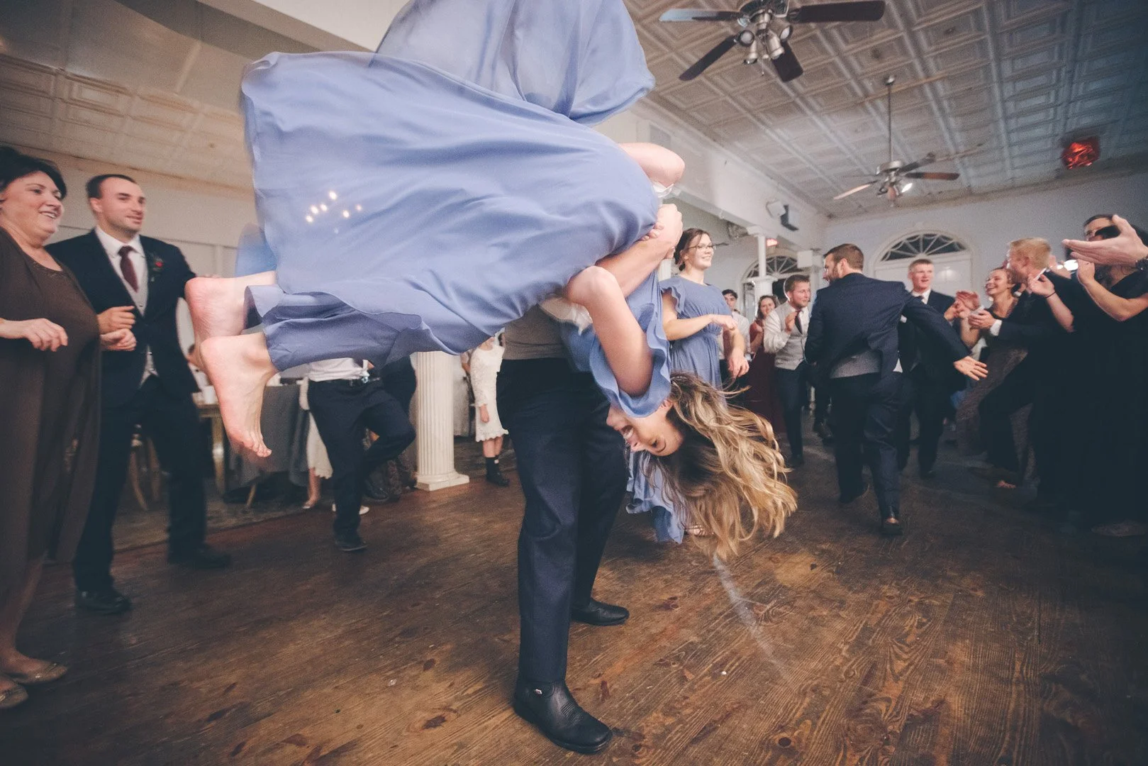 A lively dance scene at a wedding with a man in a suit lifting a laughing woman in a blue dress upside down, surrounded by cheering guests.