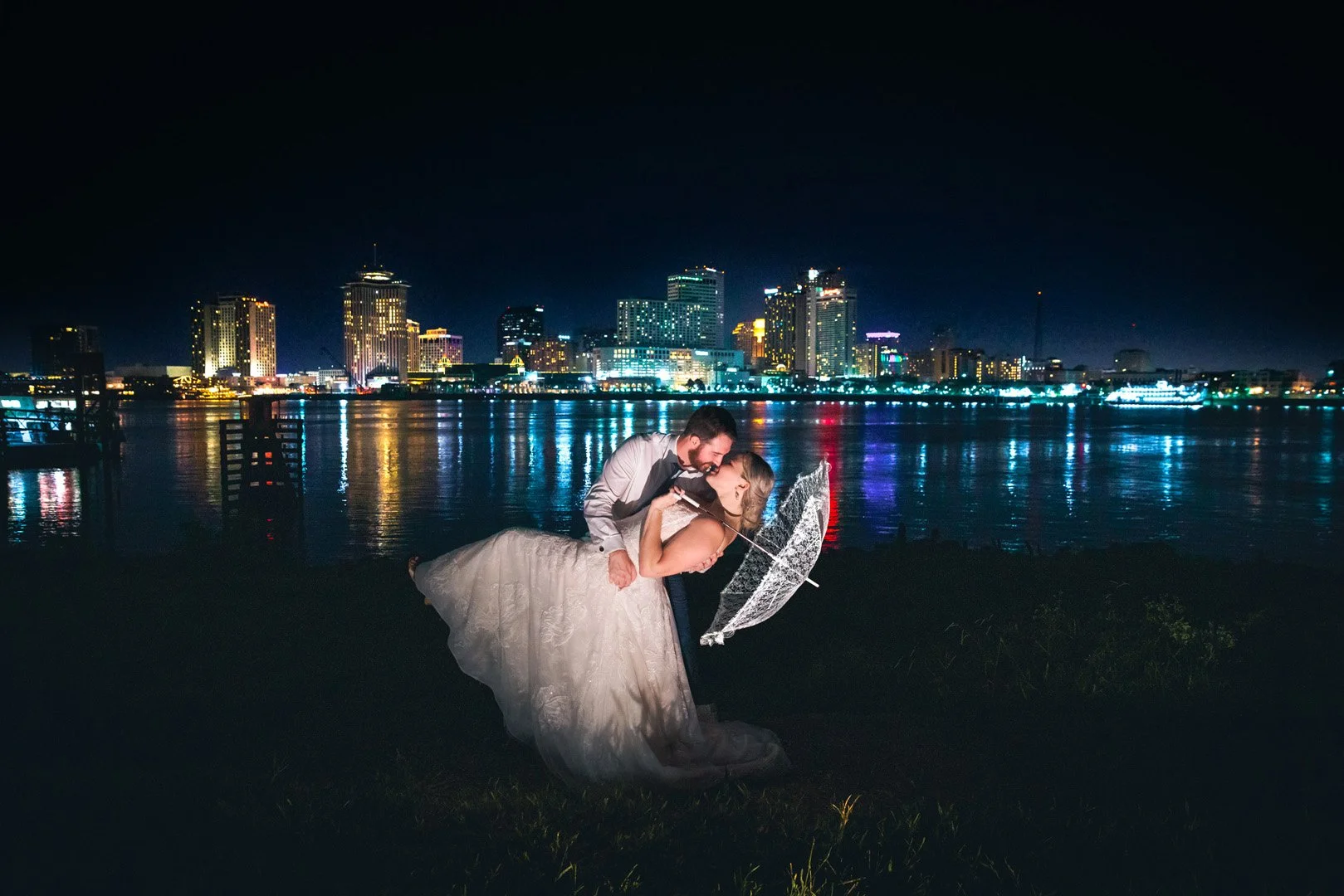 Bride and groom kissing with a lit city skyline at night in the background, next to a body of water.