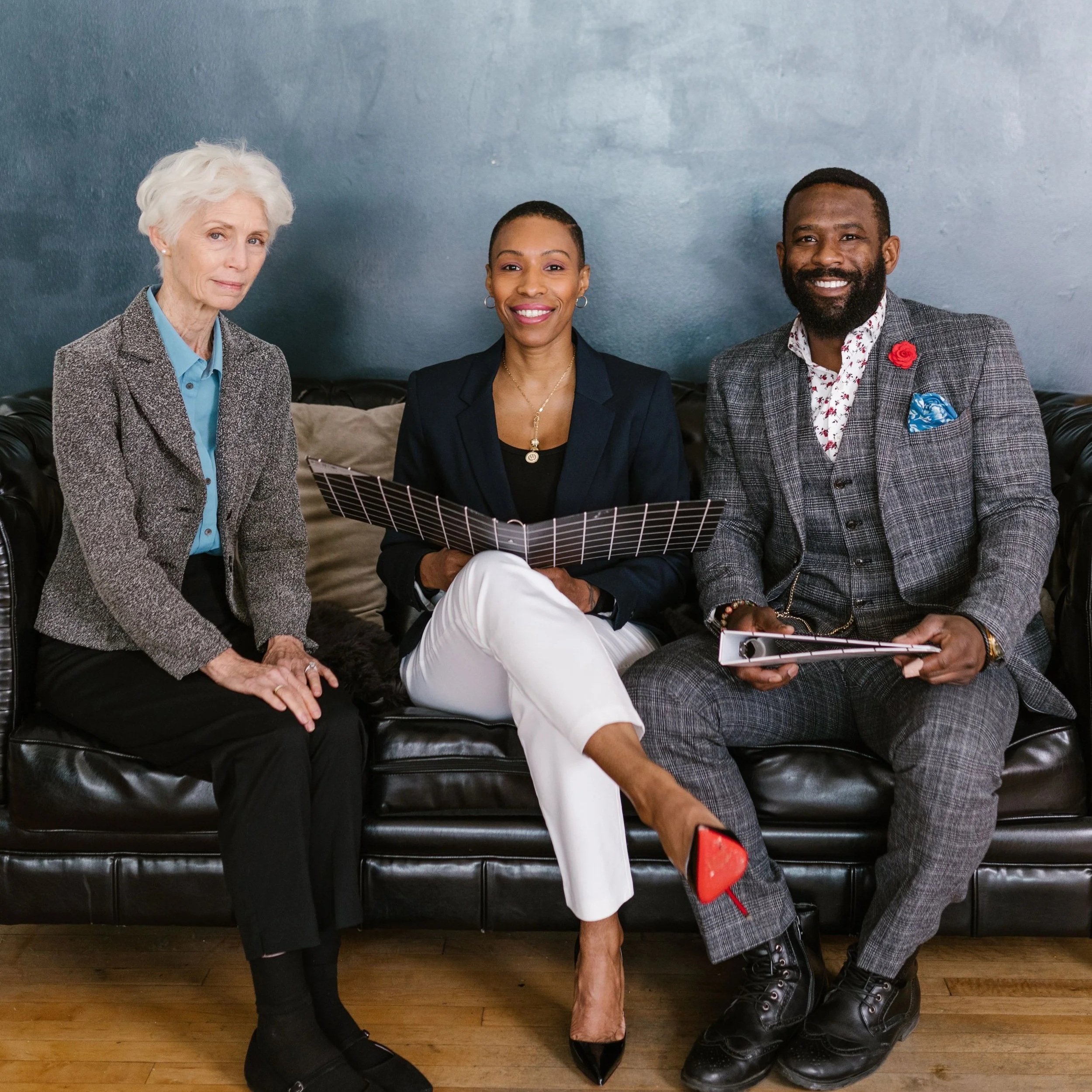 Group of three diverse people sitting on a black leather couch against a blue wall, smiling, with a laptop and tablet.