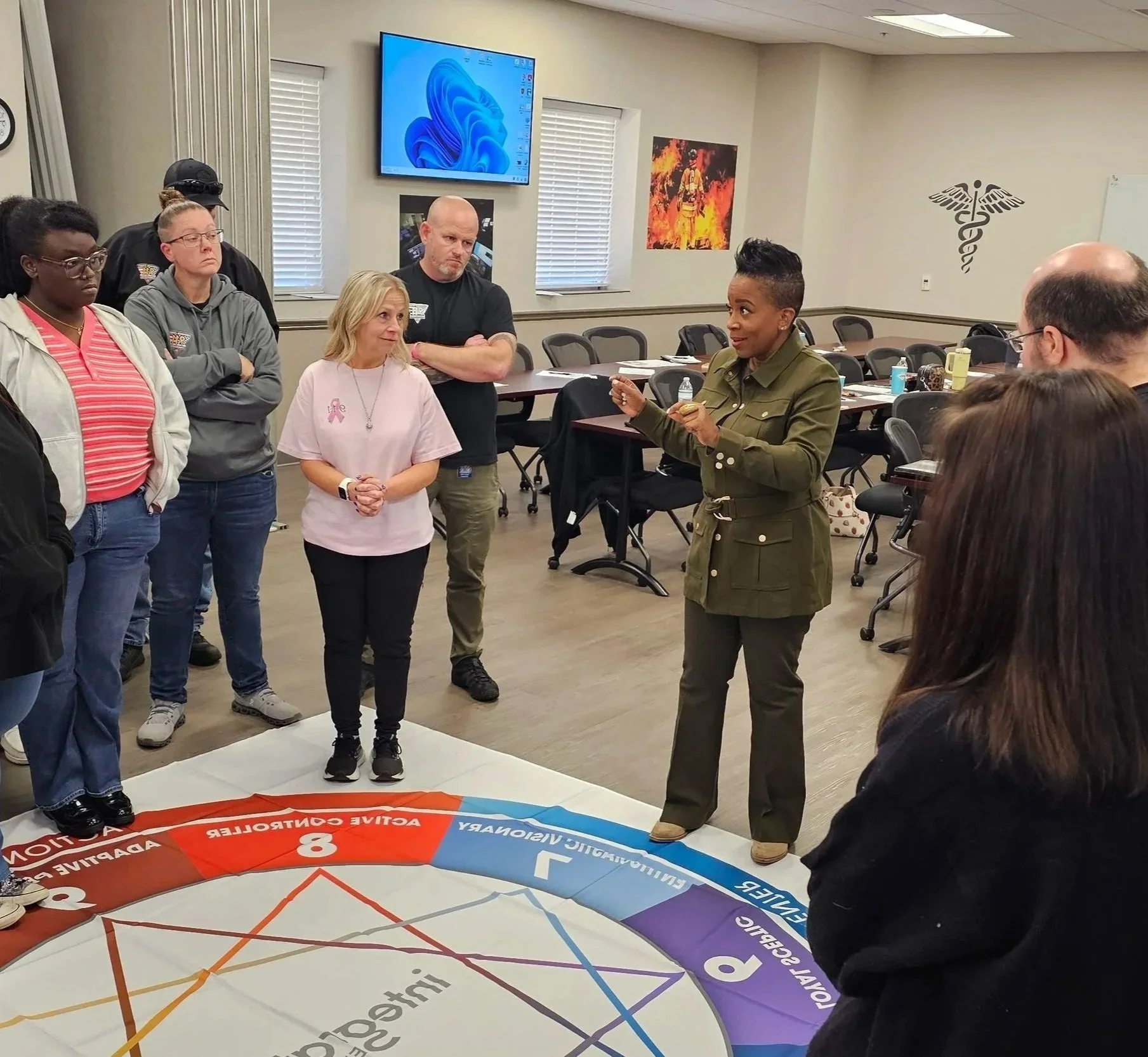 A diverse group of people standing in a room with a woman in military attire speaking. The room has a large colorful circular infographic on the floor and posters on the walls, including a firefighter and medical symbol. A large screen is mounted on the wall.