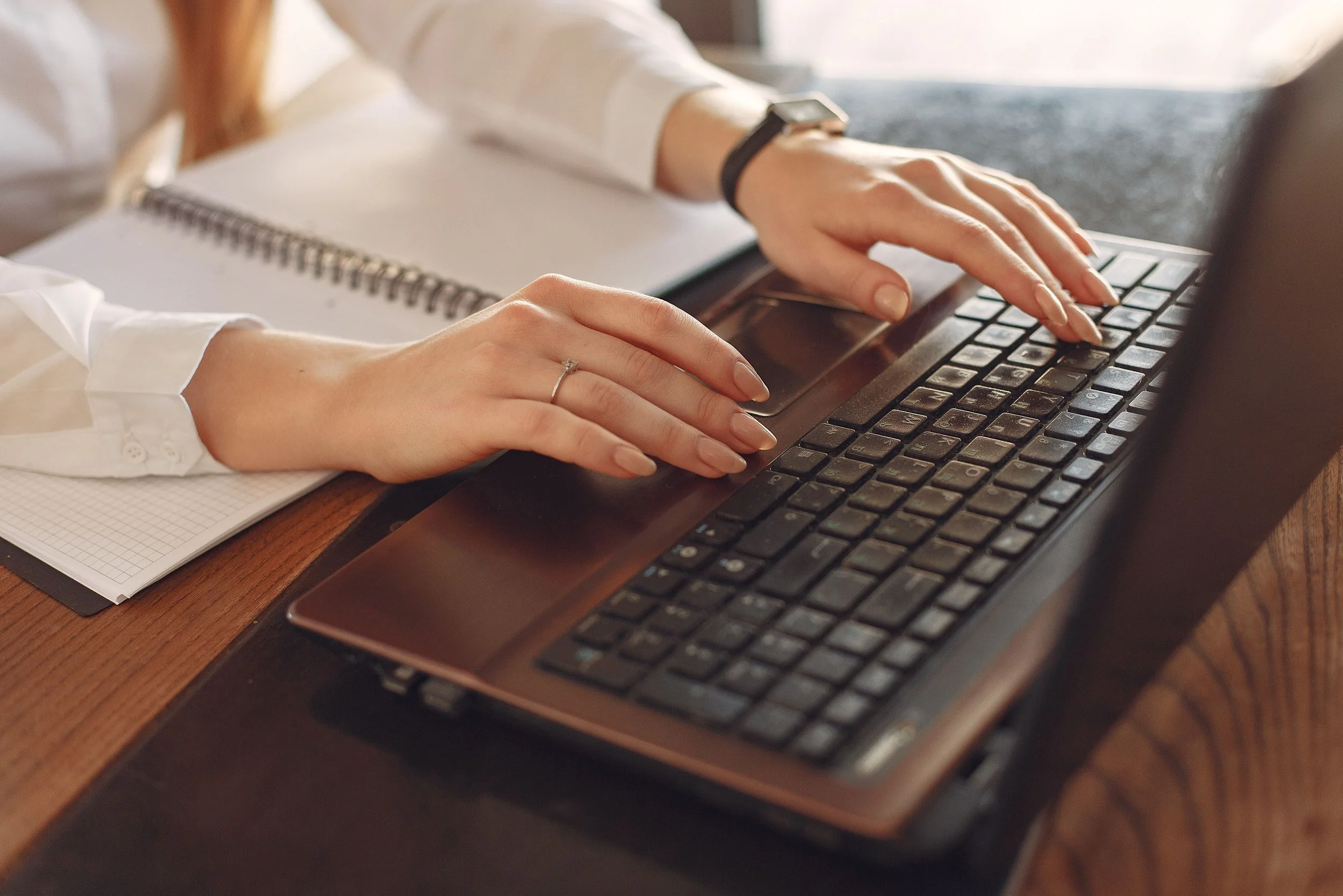 Close-up of a person typing on a laptop keyboard with a notepad and pen next to it.