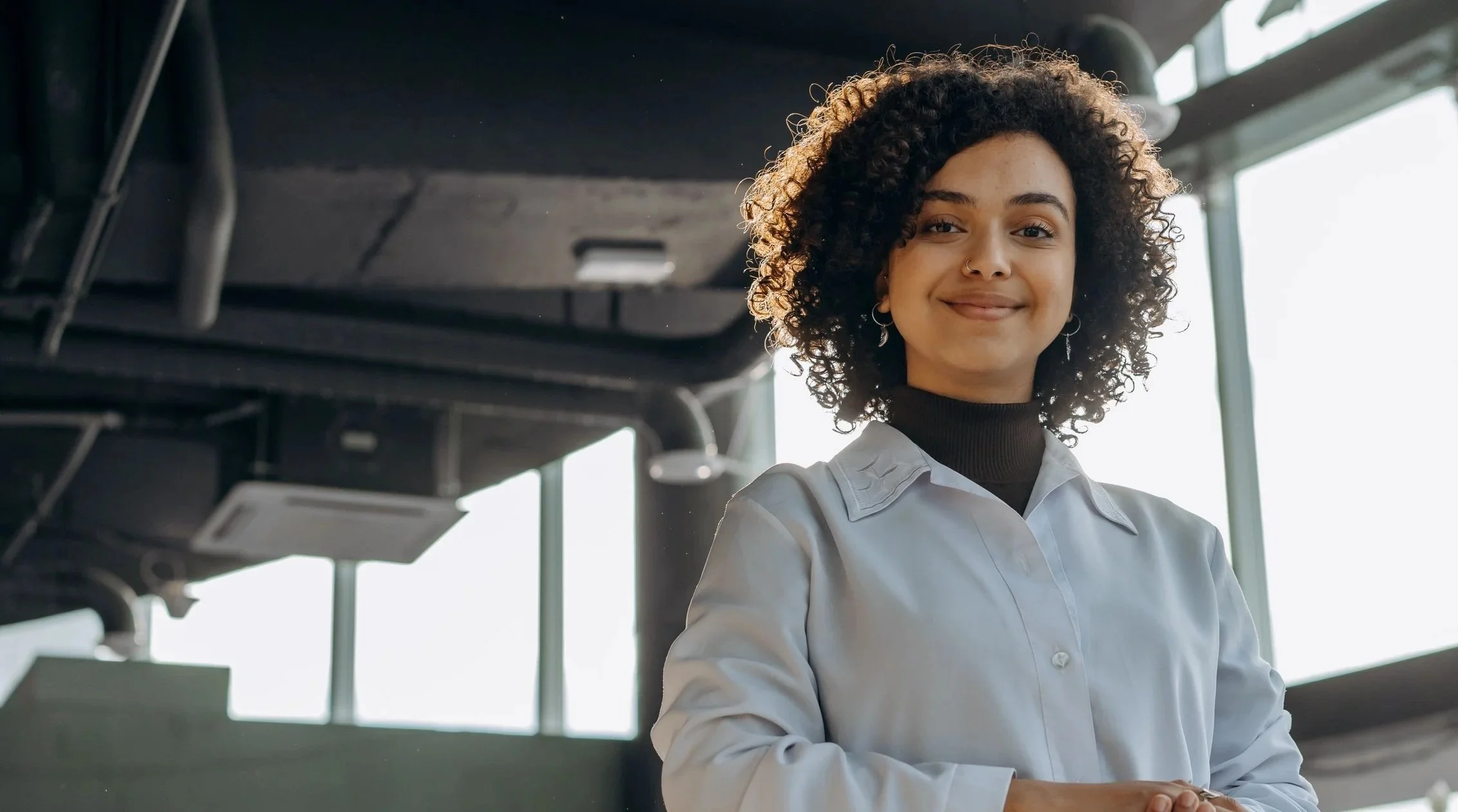 A young woman with curly hair and earrings, wearing a light-colored shirt over a black turtleneck, smiling in an indoor space with large windows.