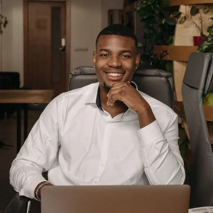 A young man in a white shirt sitting at a desk, smiling with his hand on his chin in an office setting.