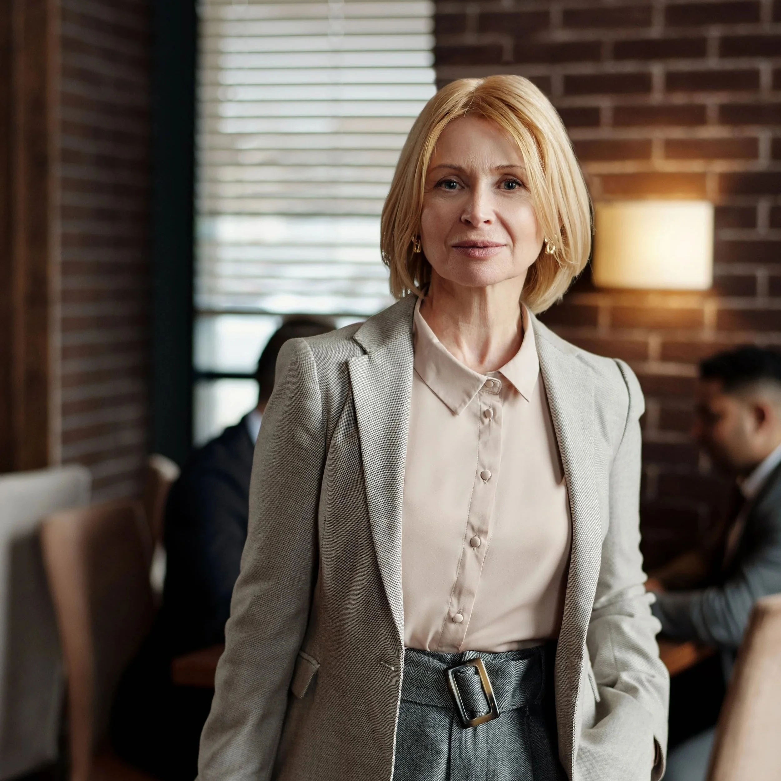 Portrait of a middle-aged woman with blonde hair, wearing a light grey blazer and a white blouse, standing in a room with a wooden wall and window blinds in the background.