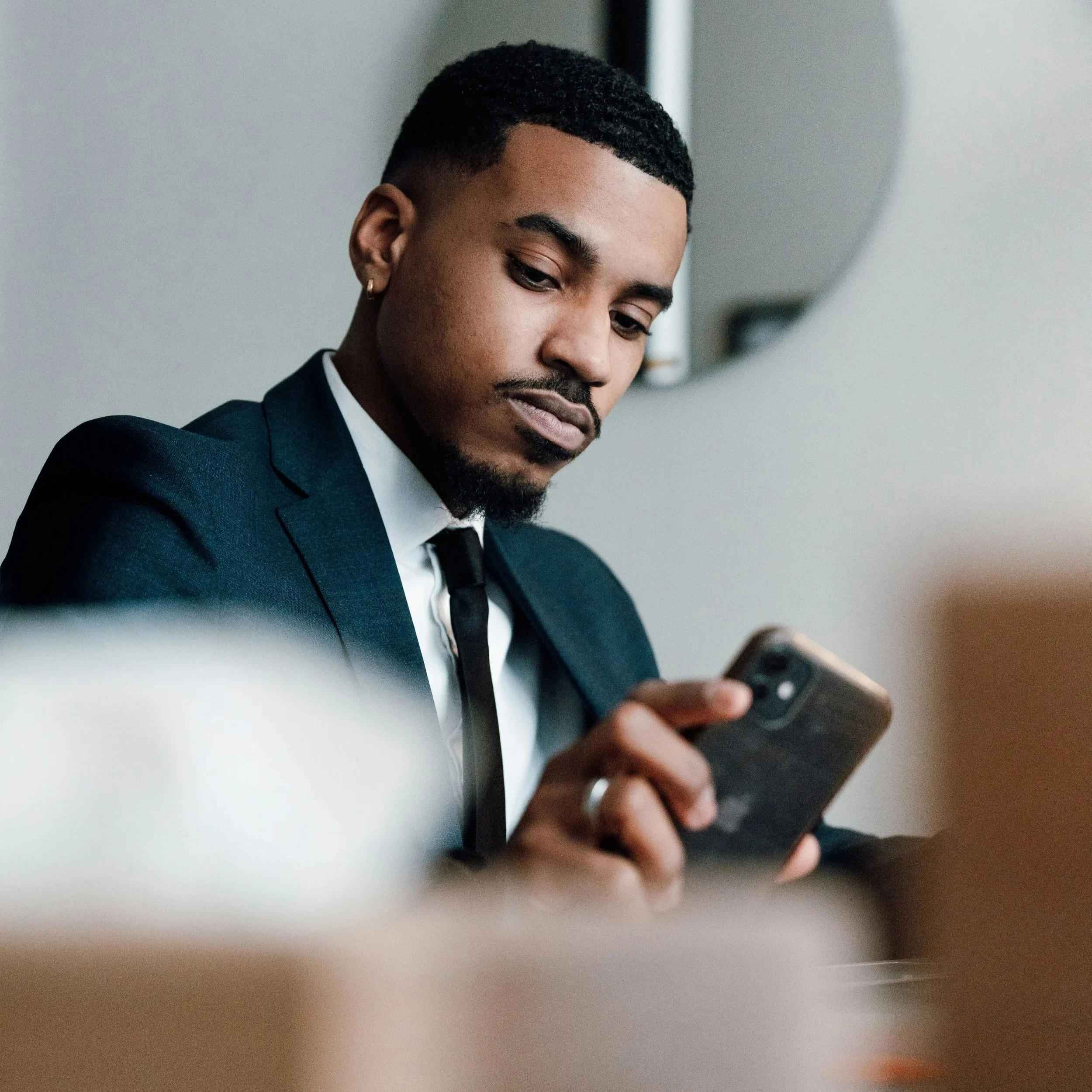 A young man with dark hair and a beard wearing a blue suit and white shirt, looking down at a phone in his hands.